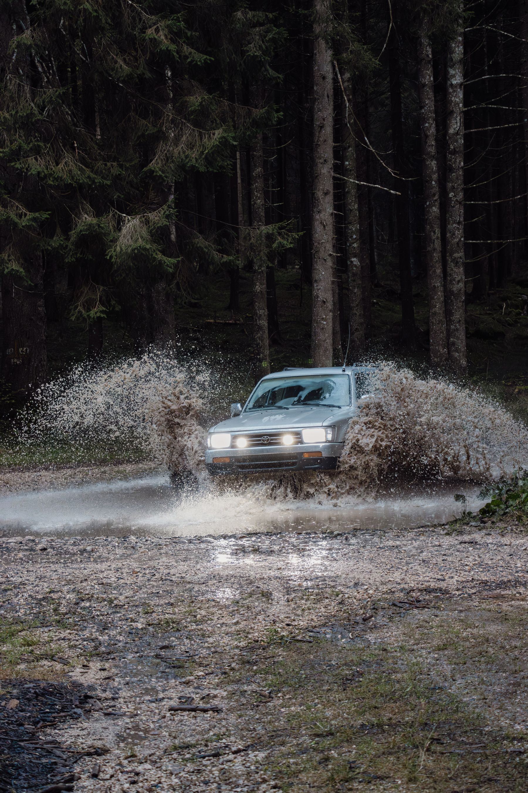 Silver second-generation 4Runner throwing up a wall of muddy water crossing a puddle in a dark pine forest