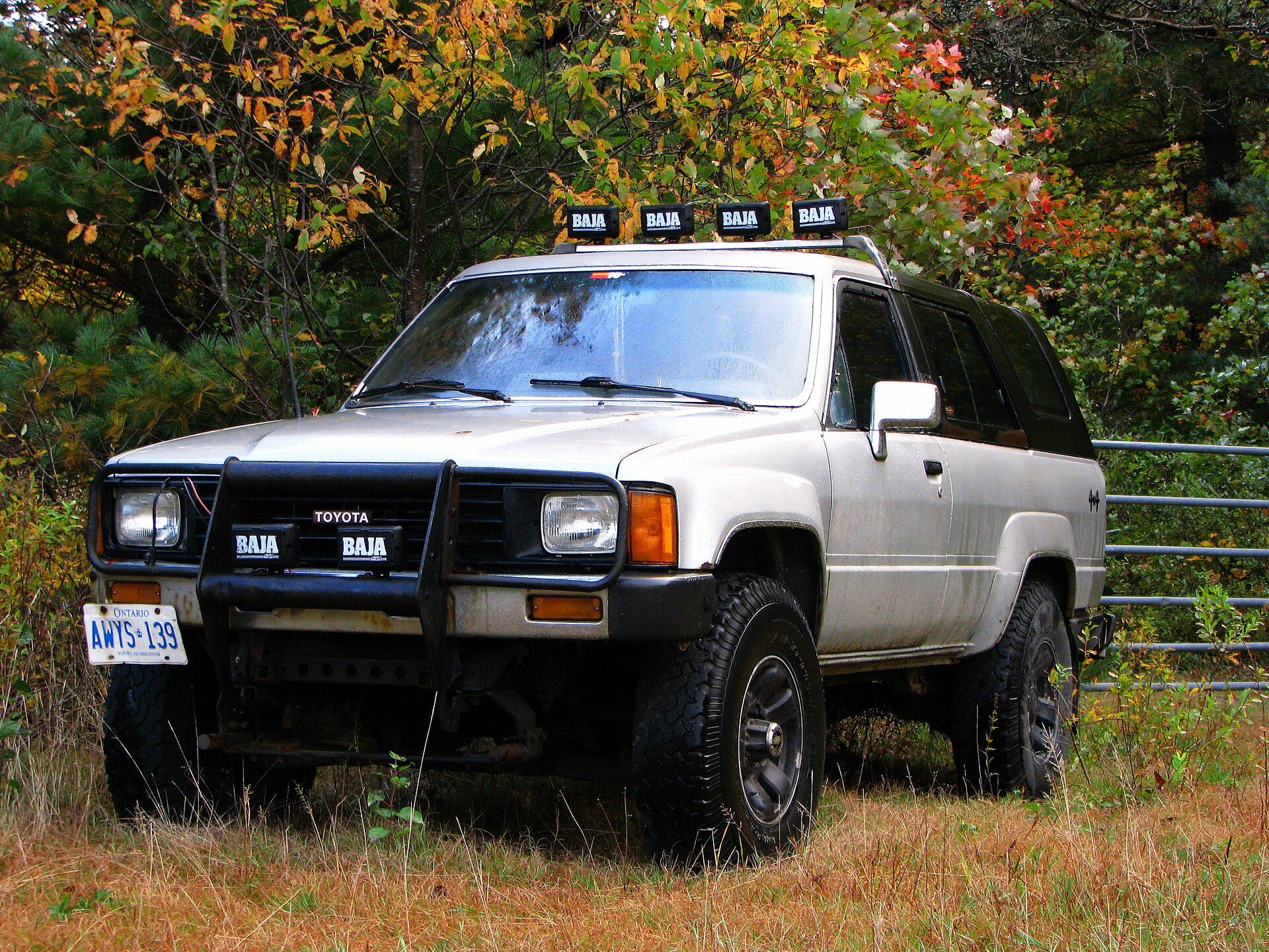 Silver first-generation two-door 4Runner with a black brush guard and roof-mounted Baja lights parked in tall grass below autumn trees