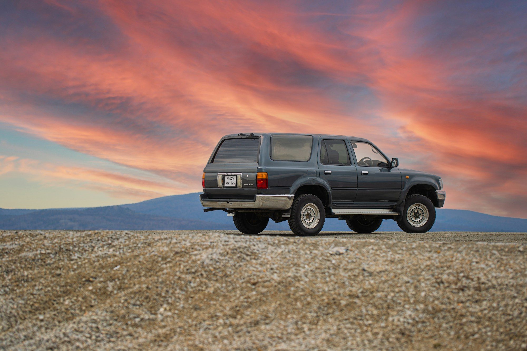 Grey second-generation Toyota Hilux Surf, the Japanese-market 4Runner, parked on a gravel rise under a red sunset sky with hills behind