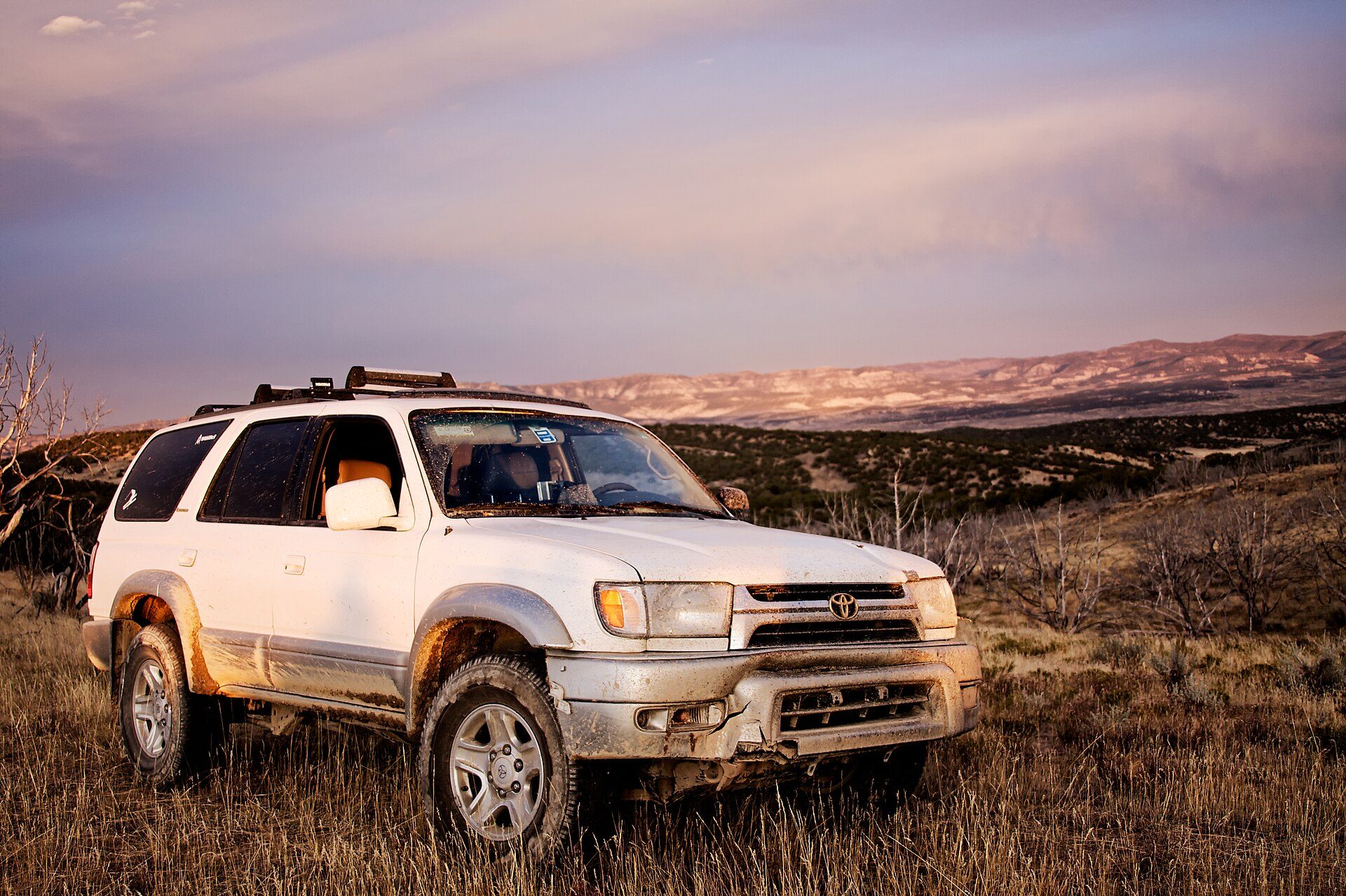 Mud-spattered white facelifted third-generation 4Runner parked in dry grass on a high-desert ridge at sunset