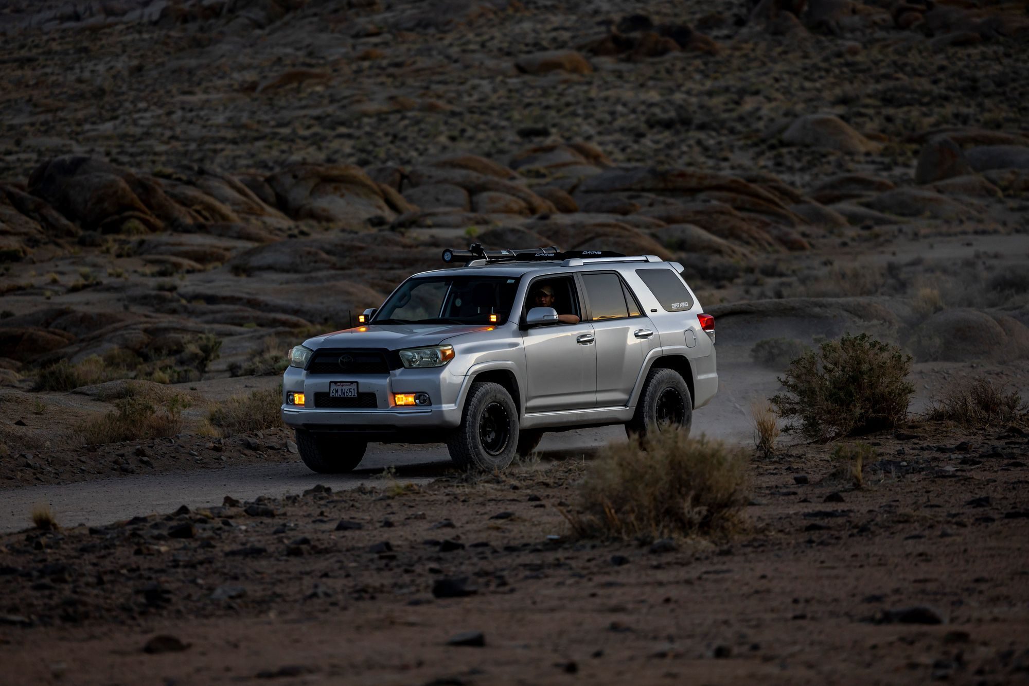 Silver early fifth-generation 4Runner with a roof rack kicking up dust on a desert track among granite boulders at dusk