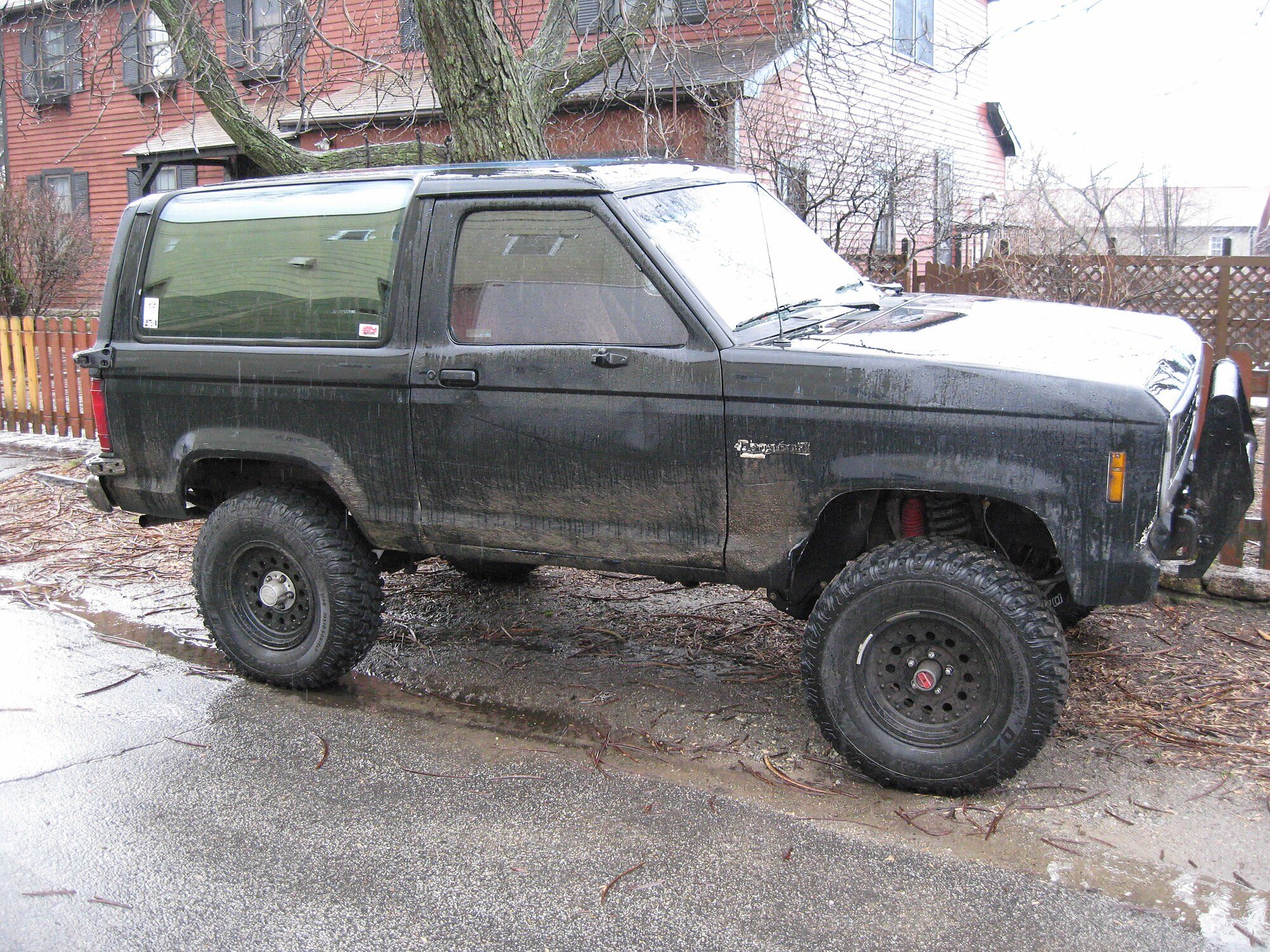 A lifted black 1984–88 Ford Bronco II caked in dried mud, on mud-terrain tires and black steel wheels, with front coil springs visible