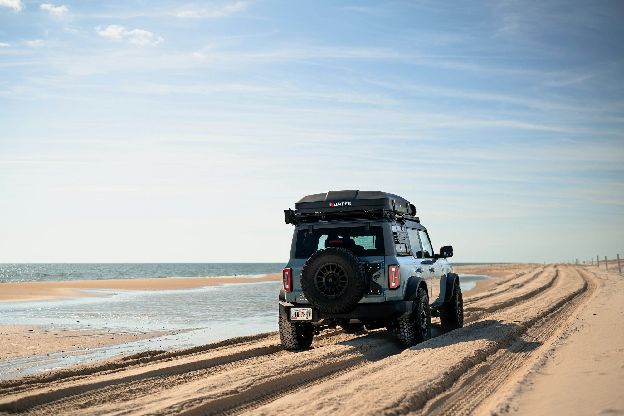 A gray four-door Ford Bronco with a rooftop tent driving away along rutted sand tracks beside the sea