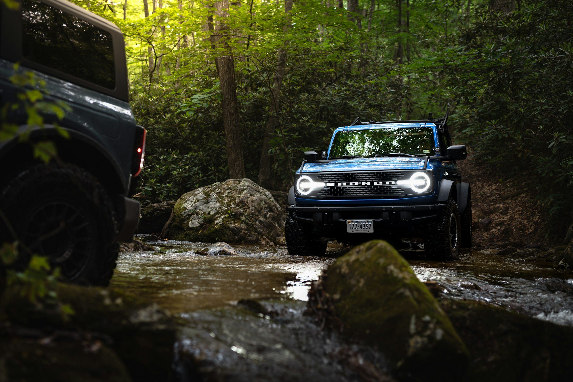 A blue sixth-generation Ford Bronco with its headlights on crossing a rocky creek in a dense forest, a second Bronco in the foreground