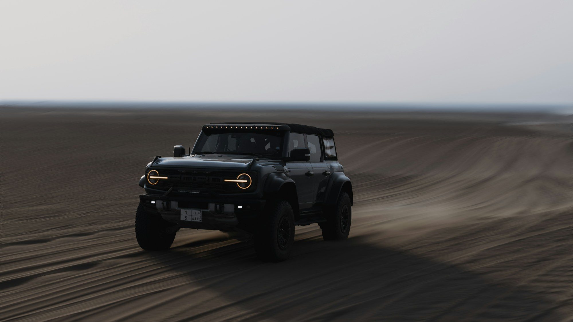 A dark Ford Bronco Raptor with amber running lights driving fast across rippled desert sand