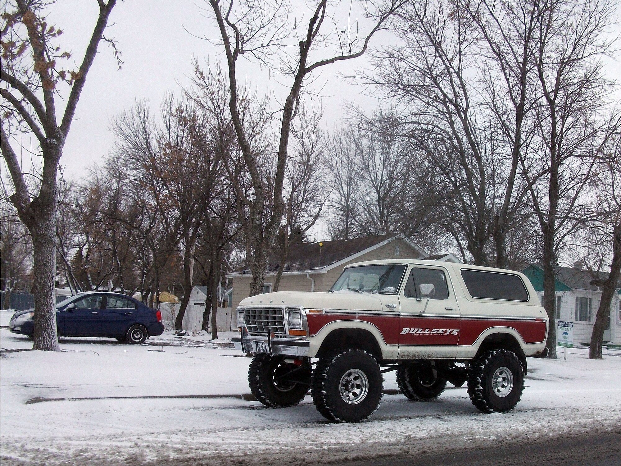 A lifted red-and-white 1978–79 Ford Bronco on big tires parked on a snowy roadside under bare trees
