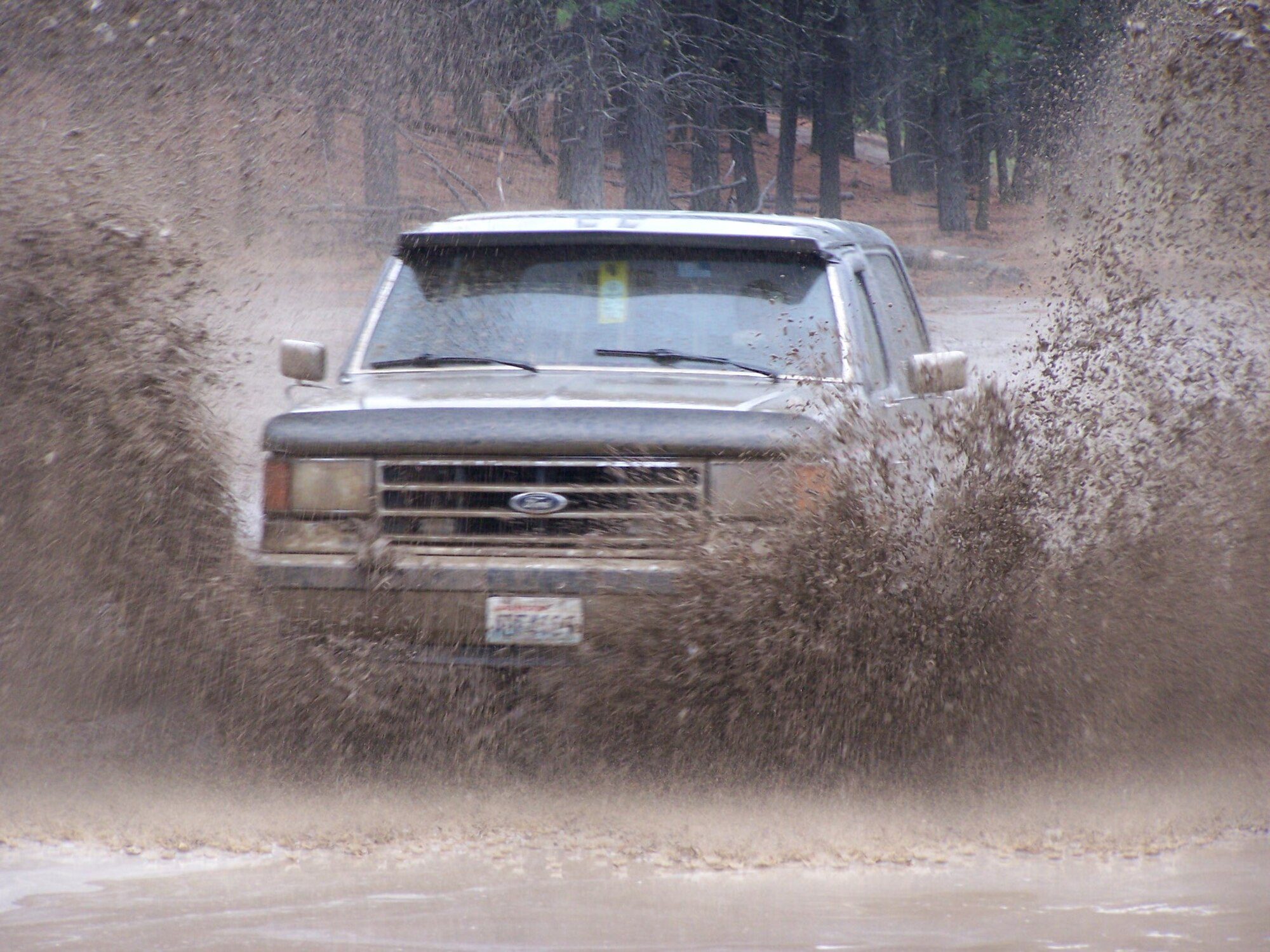 A silver 1989 Ford Bronco throwing up a wall of mud as it drives through a flooded track among pine trees