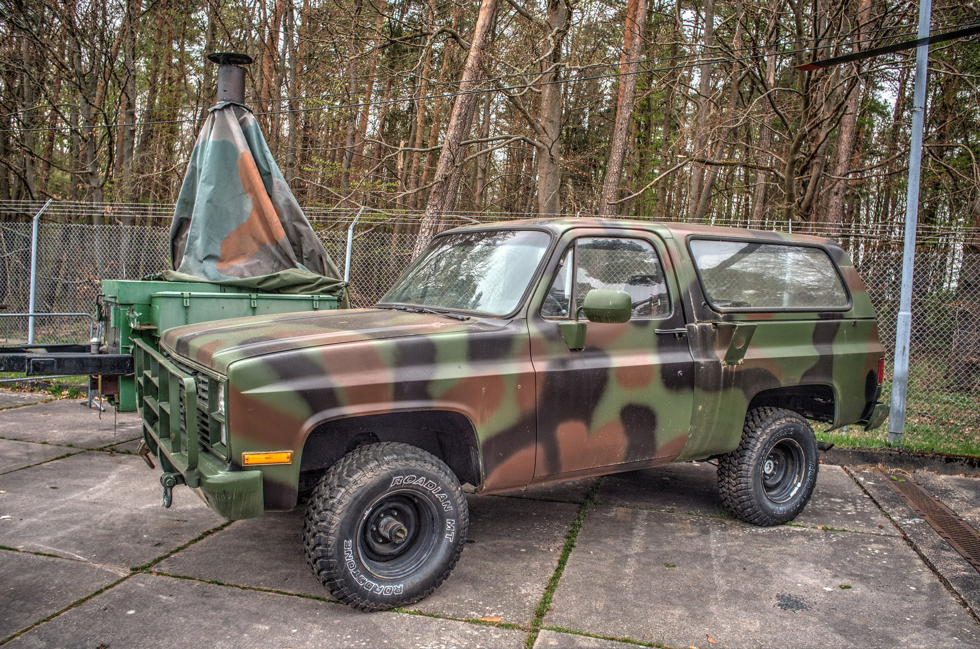 A woodland-camouflage M1009 CUCV K5 Blazer with brush guard and mud tires, next to a camouflaged trailer among pine trees