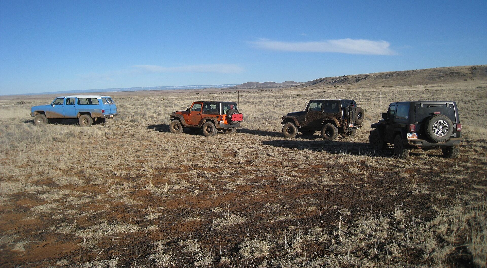A blue and white square-body Suburban with mud-caked sides leads three Jeep Wranglers across open grassland on a 4x4 club run near Hat Knoll