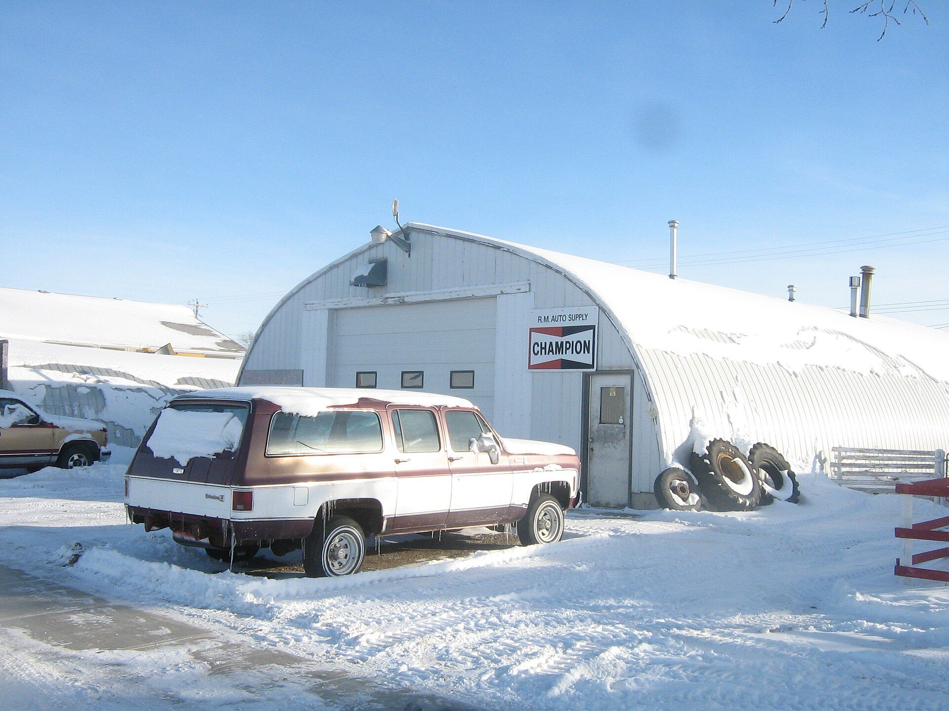 A two-tone 1973–80 Suburban Silverado with a tailgate parked in deep snow beside a Quonset-hut auto supply