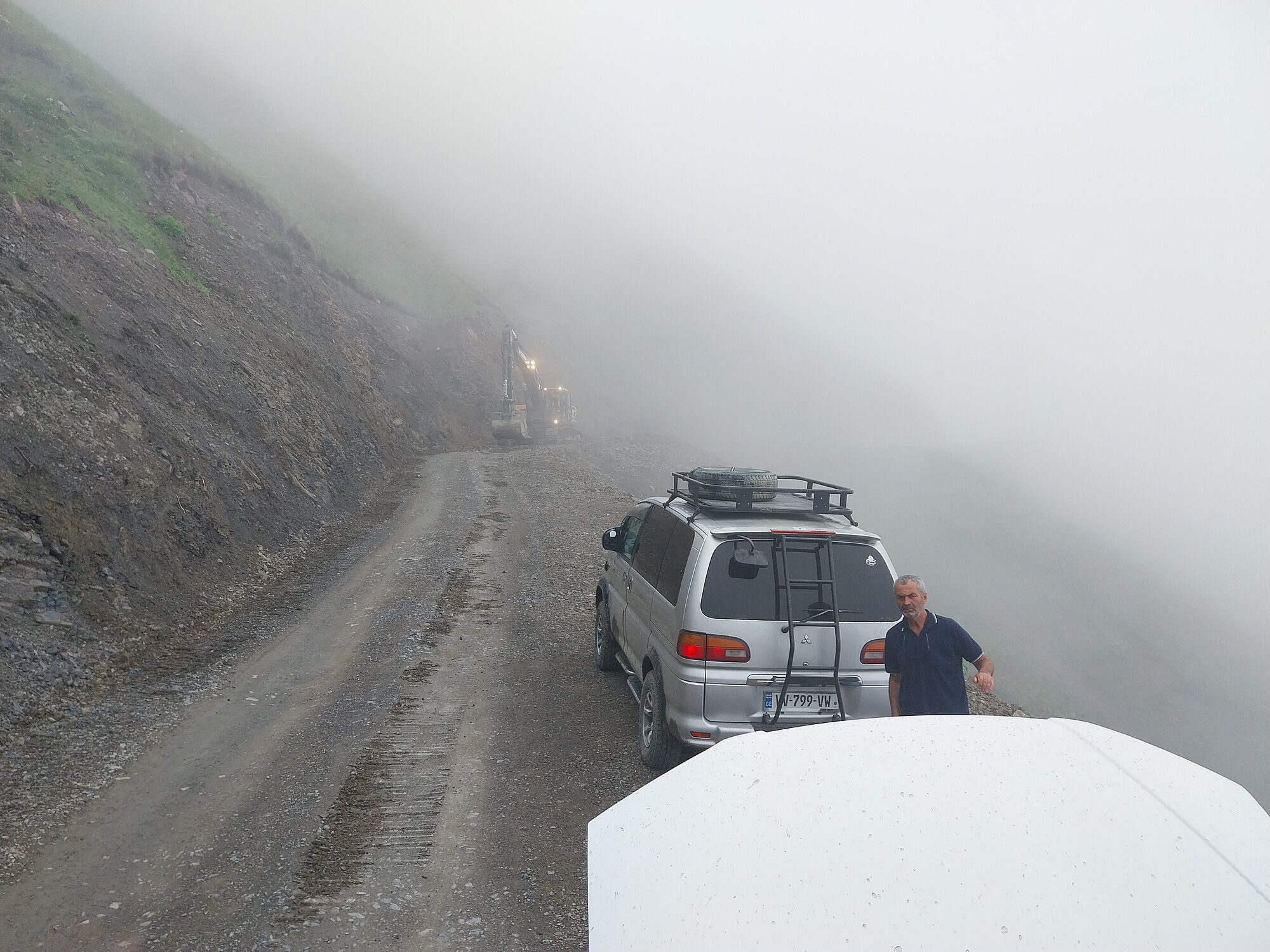 A silver Delica Space Gear with a roof rack and rear ladder on a narrow gravel shelf road in the clouds below Abano Pass