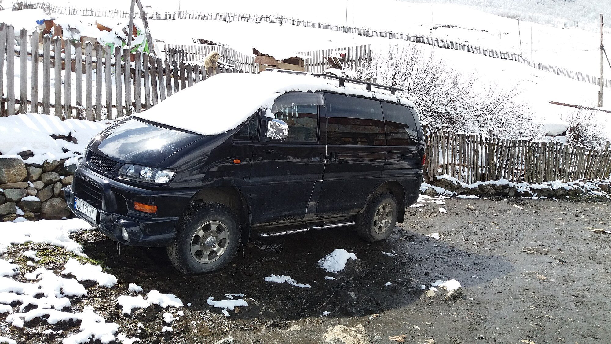 A dark blue Series 2 Delica Space Gear with snow on its roof, parked by a wooden fence in the mountain village of Ushguli
