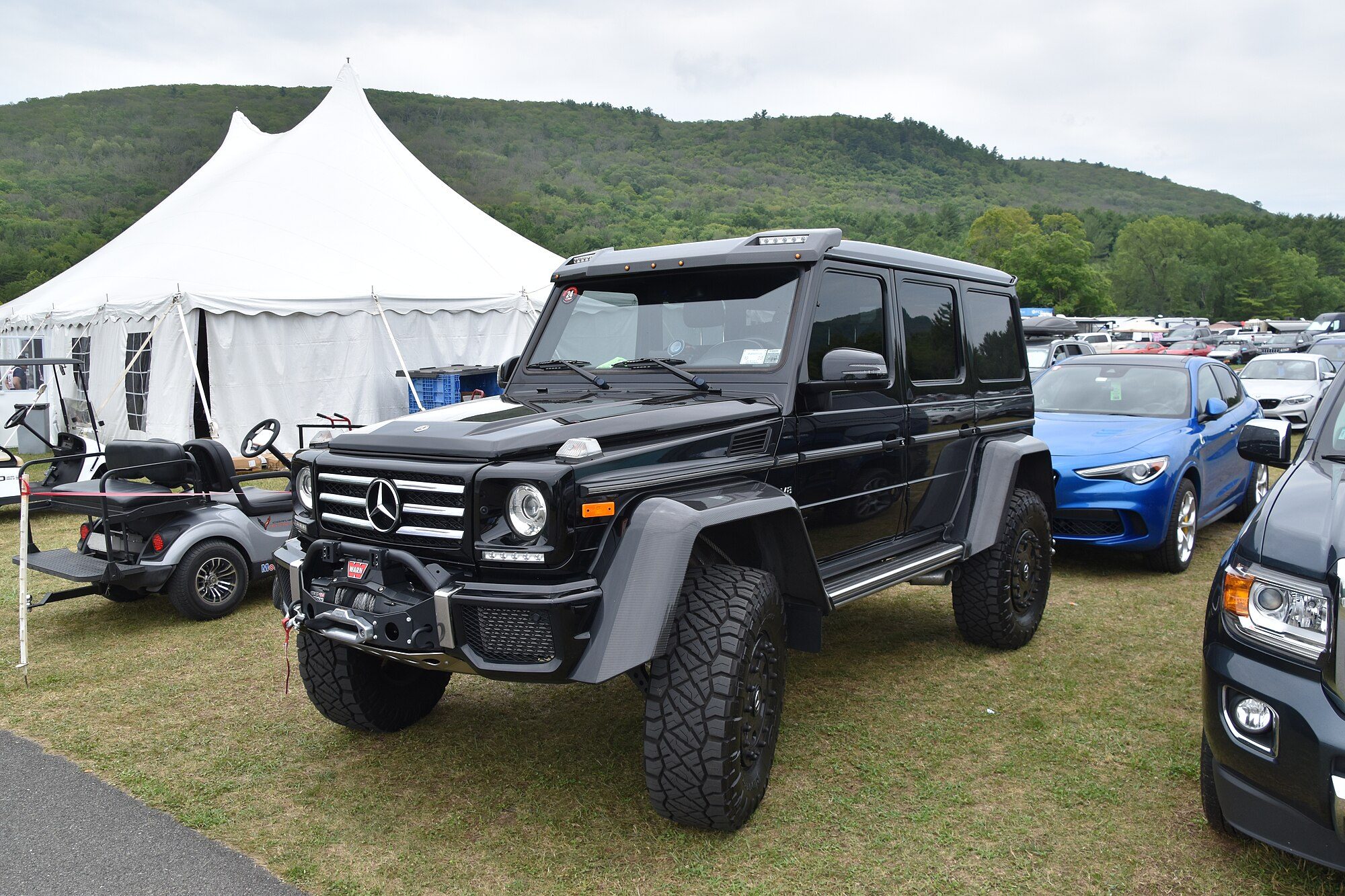 A black G 500 4×4² with an aftermarket Warn winch bumper, roof lights and all-terrain tires on black wheels, parked on a car-show field below wooded hills