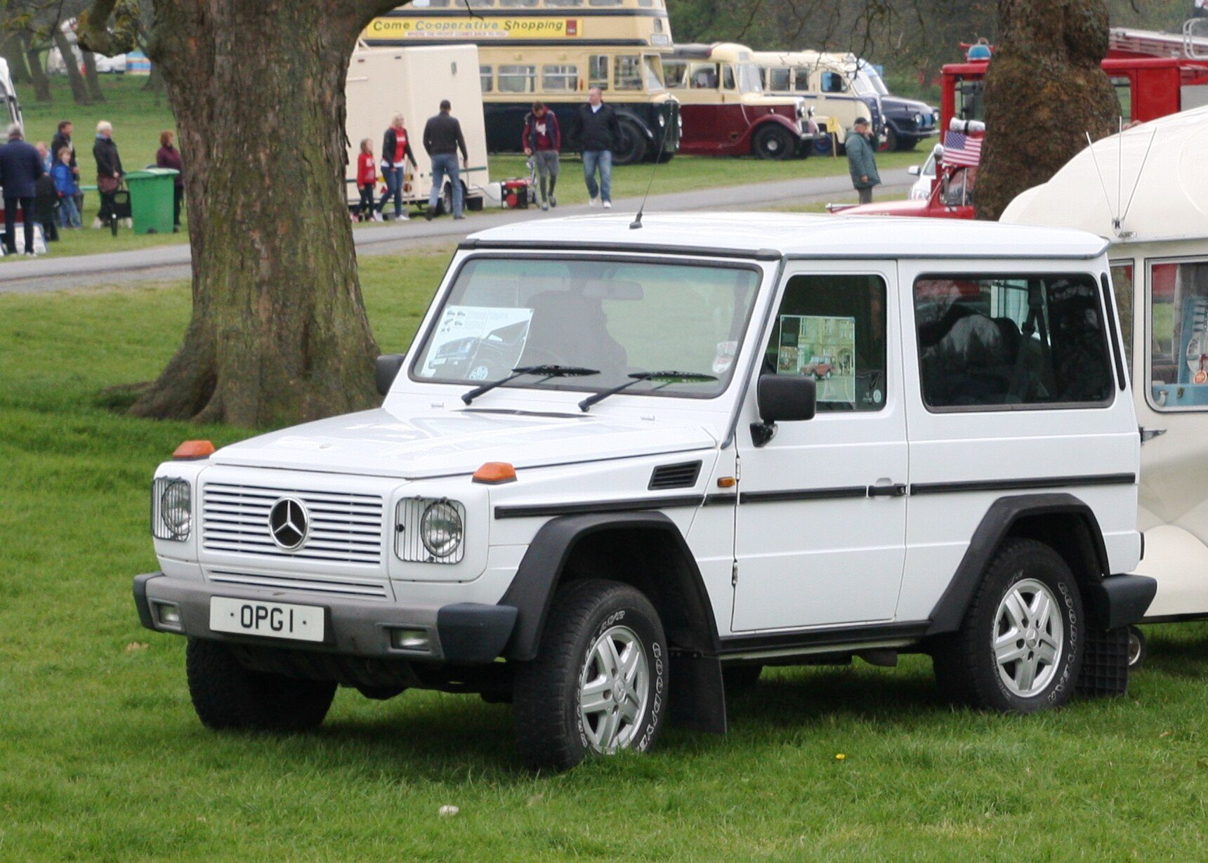 A white UK-registered 1990s short-wheelbase W463 G-Class with orange fender-top indicators parked on grass at a show (a European-market truck, not a Europa import)