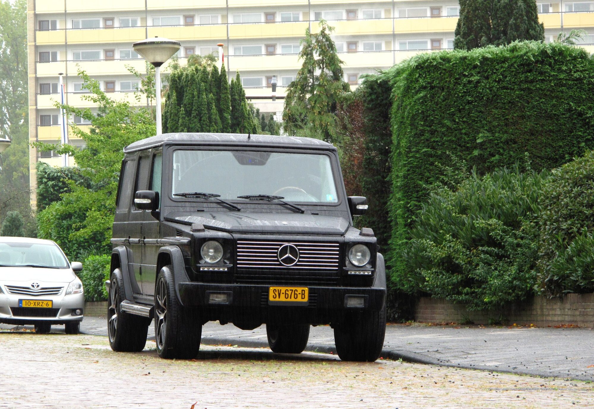 A black 2003 G 500 with Dutch number plates parked on a brick-paved street in front of hedges and an apartment block