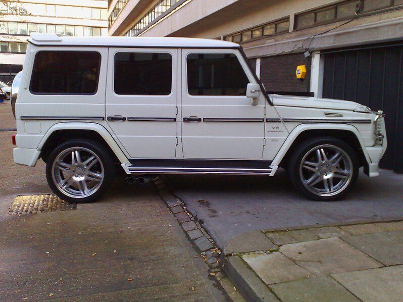 A white G 55 AMG Kompressor with V8 Kompressor wing badge and aftermarket multi-spoke wheels in side profile on a city street
