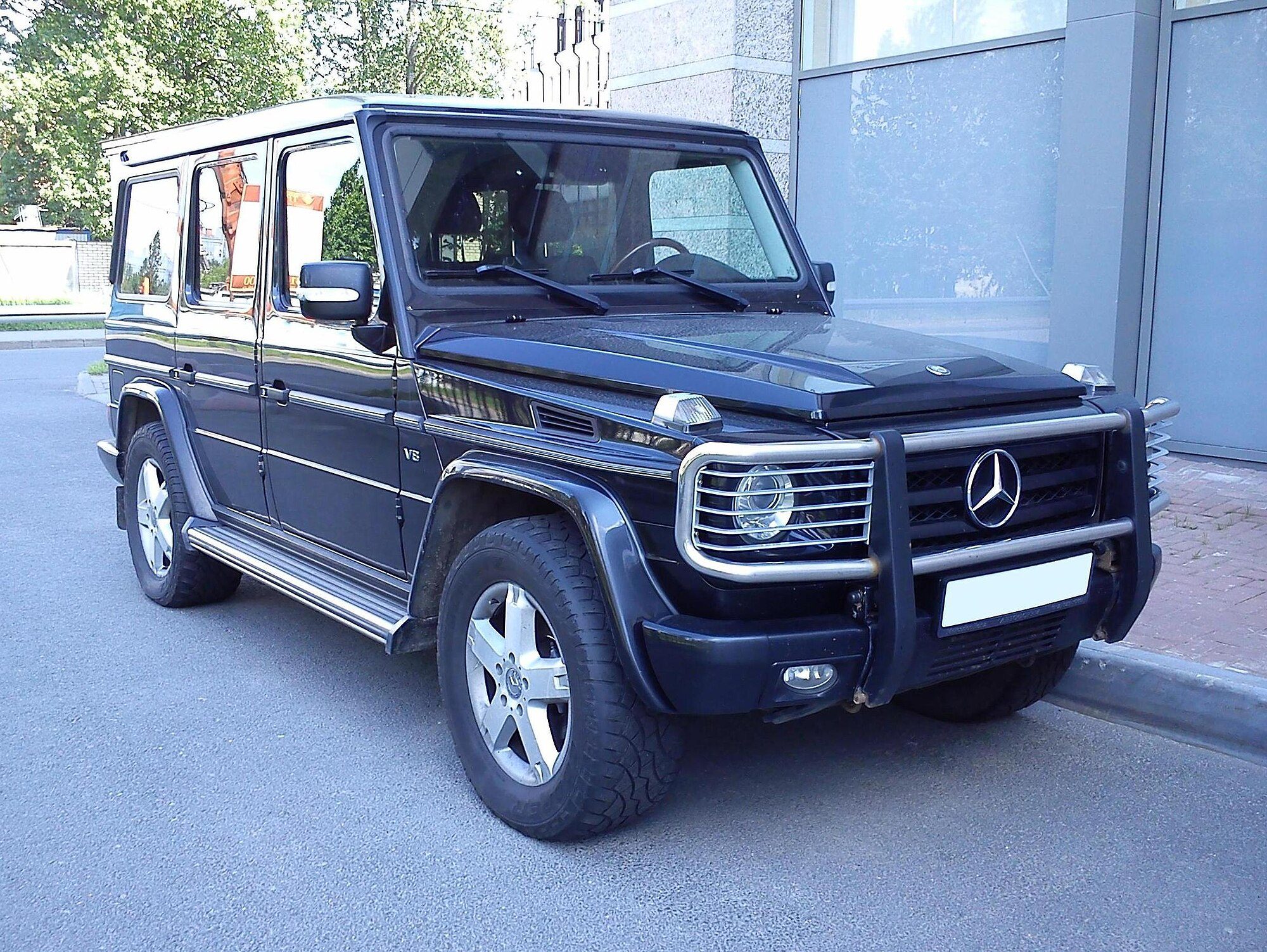 A black W463 G-Class with V8 badge, bi-xenon headlamps and a chrome brush guard parked at a kerb; no LED running lamps, so a pre-2013 truck