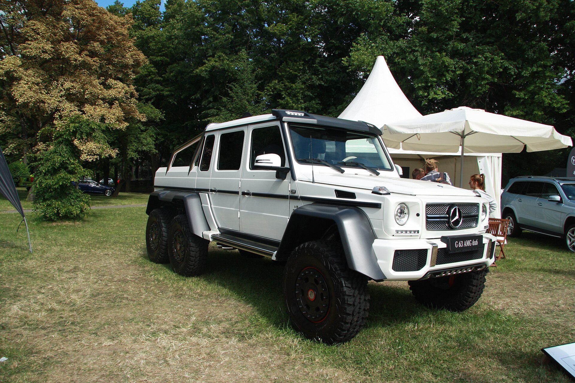 A white G 63 AMG 6×6 pickup with beadlock wheels and 37-inch tyres parked on grass at an outdoor show