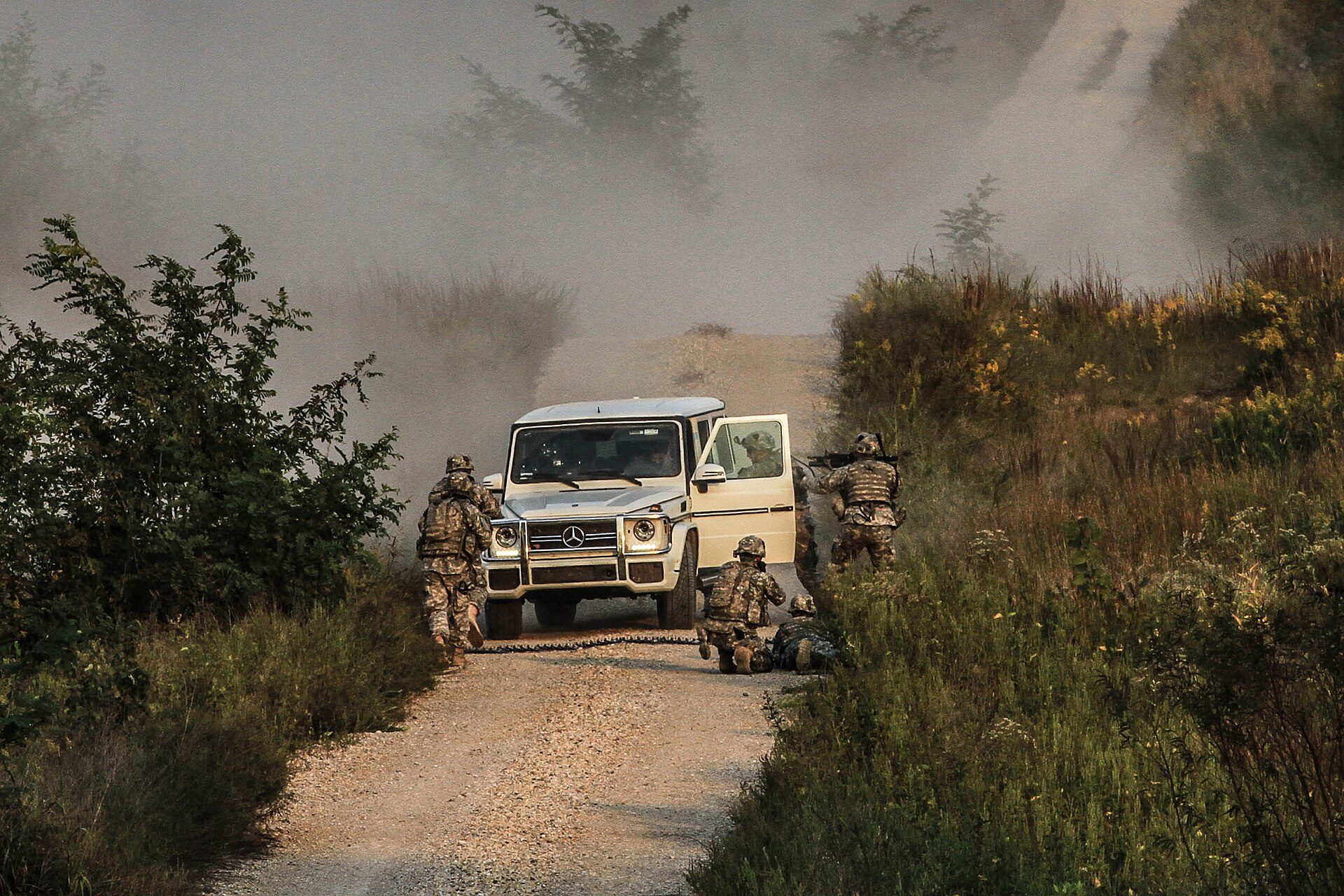 A white 2012-on G 63 AMG with LED running lights and the AMG three-intake bumper stopped on a gravel track in smoke, surrounded by soldiers during a Top Gear USA shoot at Fort Campbell
