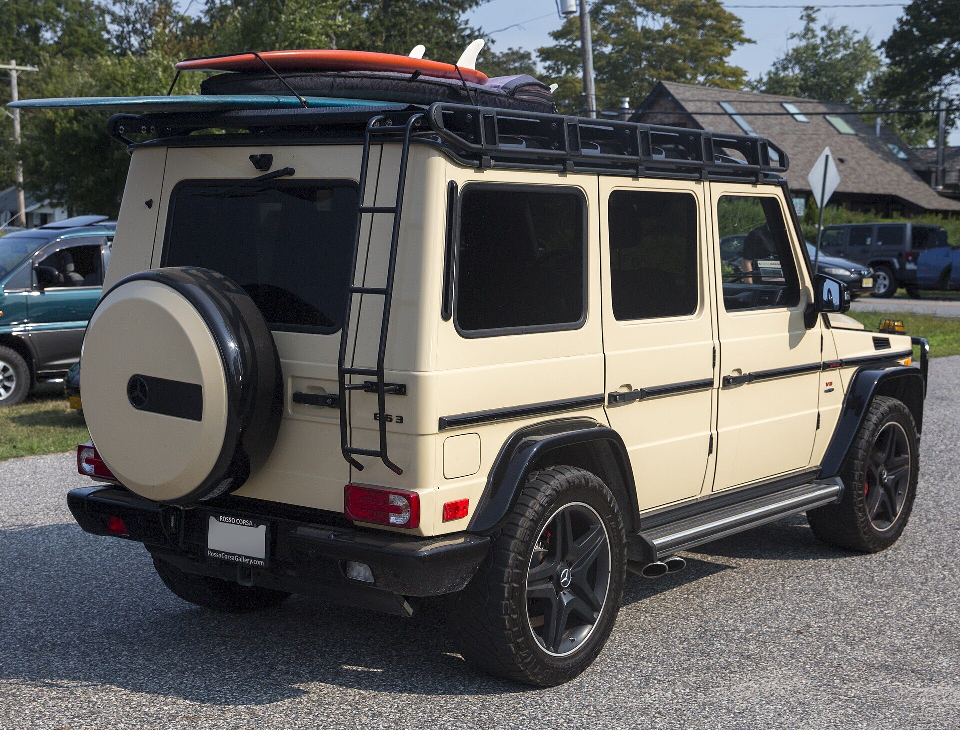 A sand-coloured 2013 G 63 AMG with a roof rack, ladder and surfboards, seen from the rear on a gravel lot