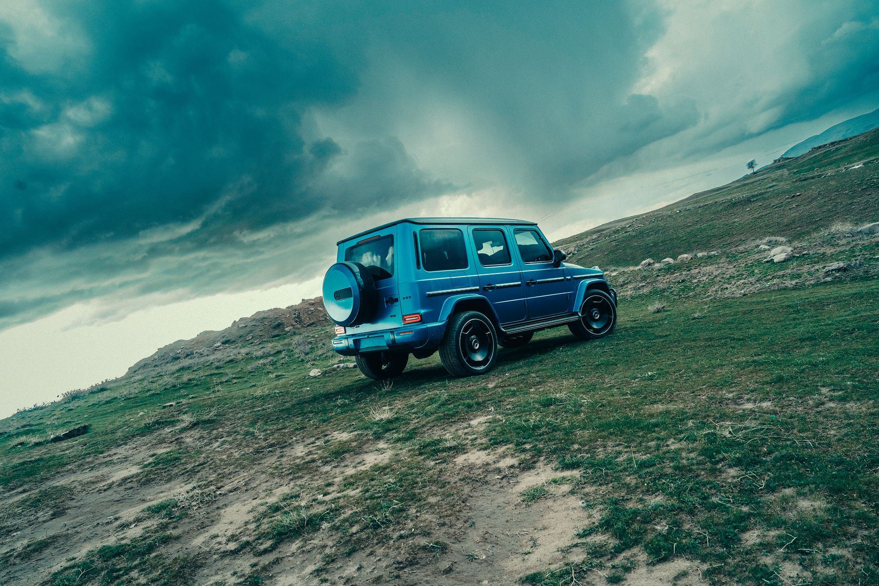 A blue second-generation Mercedes-AMG G 63, seen from the rear, on a steep grassy hillside under storm clouds
