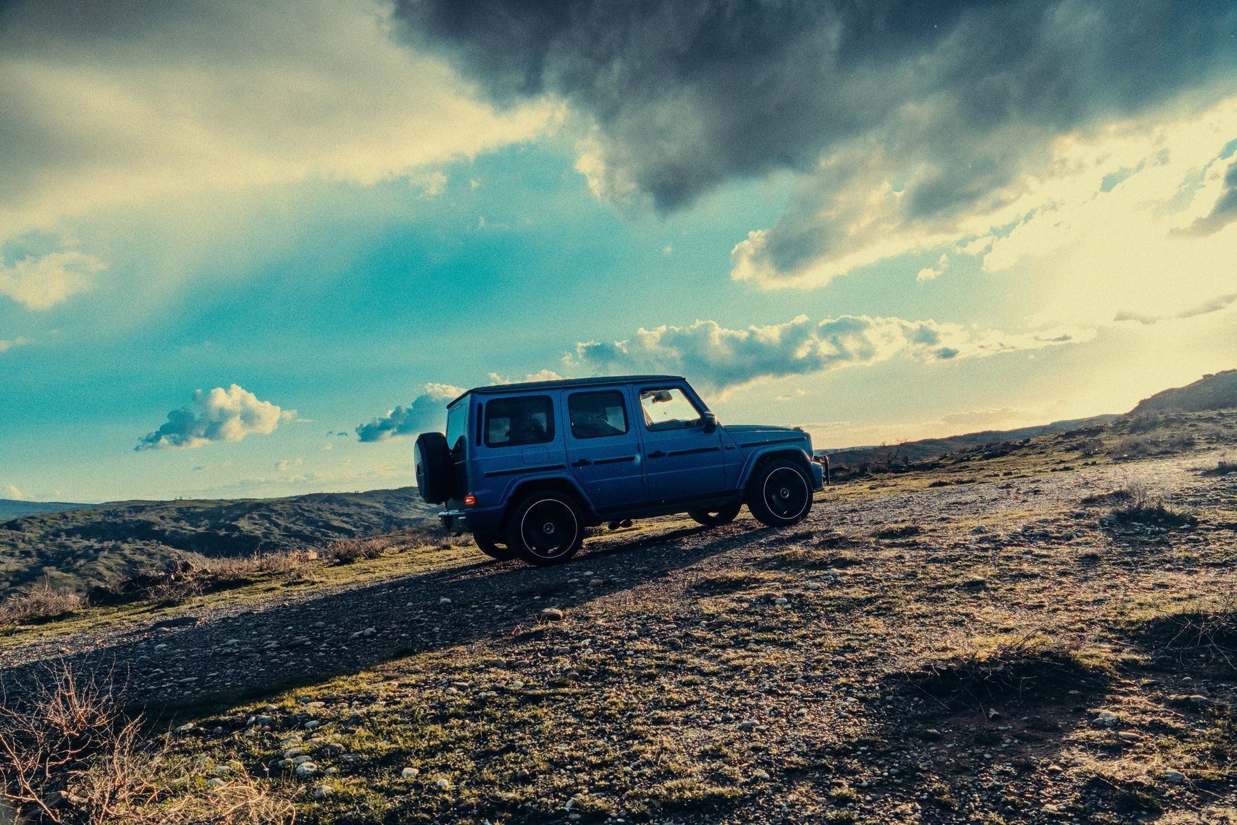 A blue second-generation Mercedes-AMG G 63 in profile on a stony ridge track at sunset
