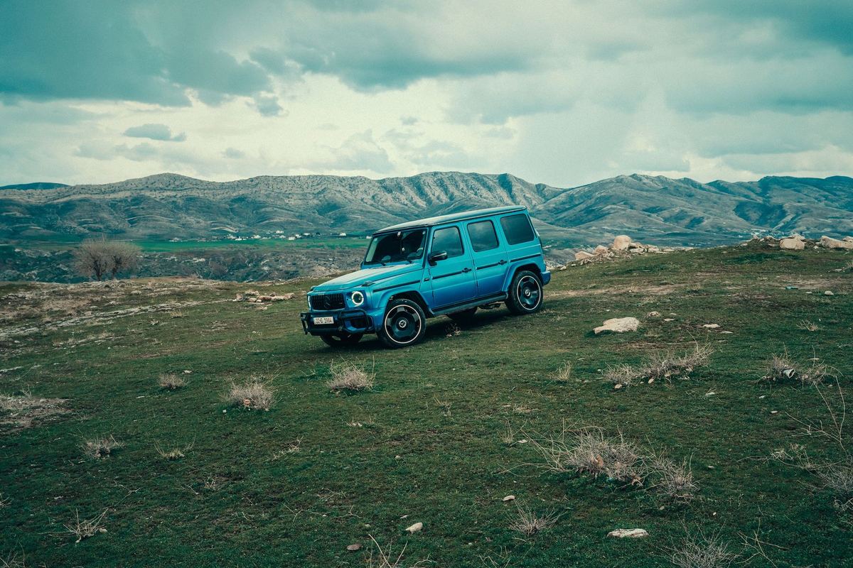 A blue second-generation Mercedes-AMG G 63 on rough grass and rocks with mountains behind