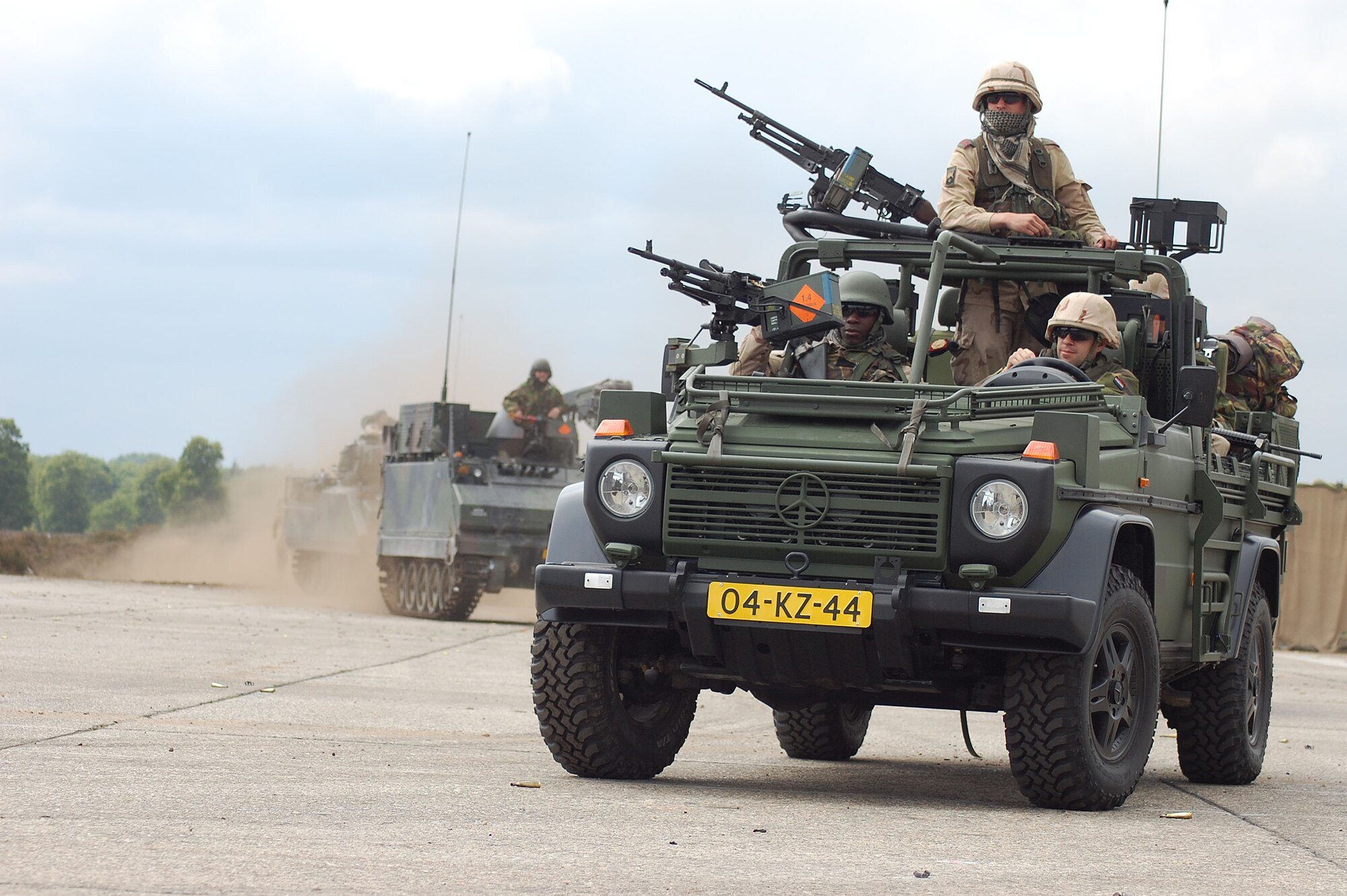 An open Dutch Army W461 290 GD weapons carrier with crew, an armoured vehicle kicking up dust behind