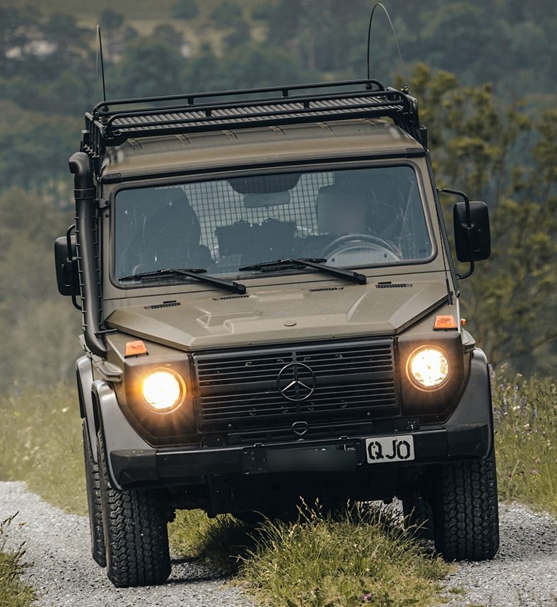 A Swiss Army W461 G 300 CDI with roof rack and snorkel nosing over rough grass and gravel, headlights on
