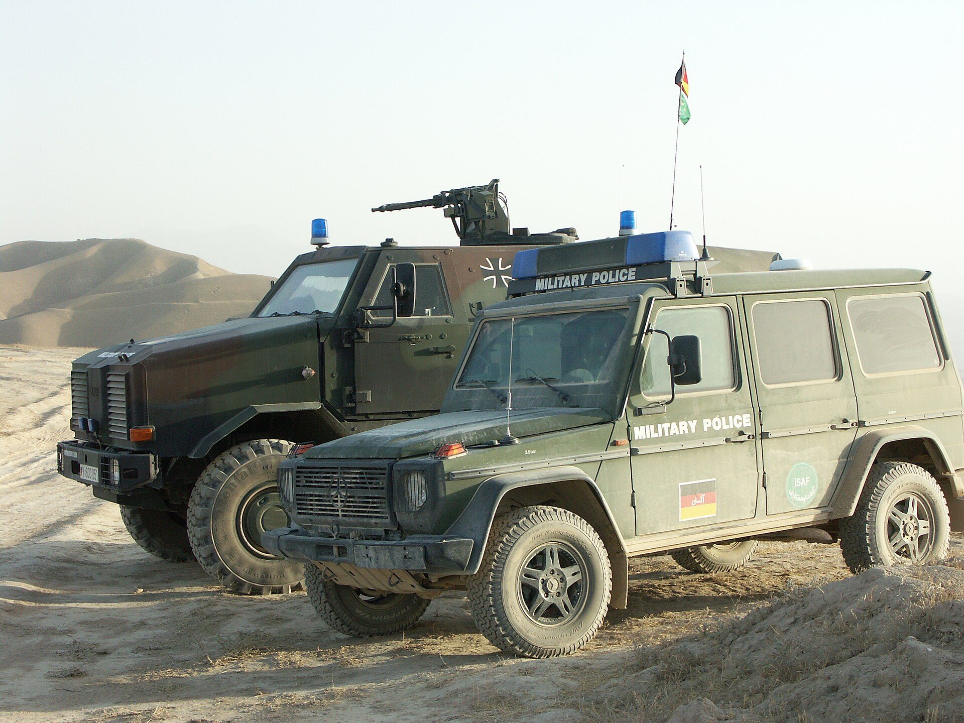 A German Army W461 "Wolf" military police wagon beside a Dingo armoured truck on a dusty plain below bare hills in Afghanistan