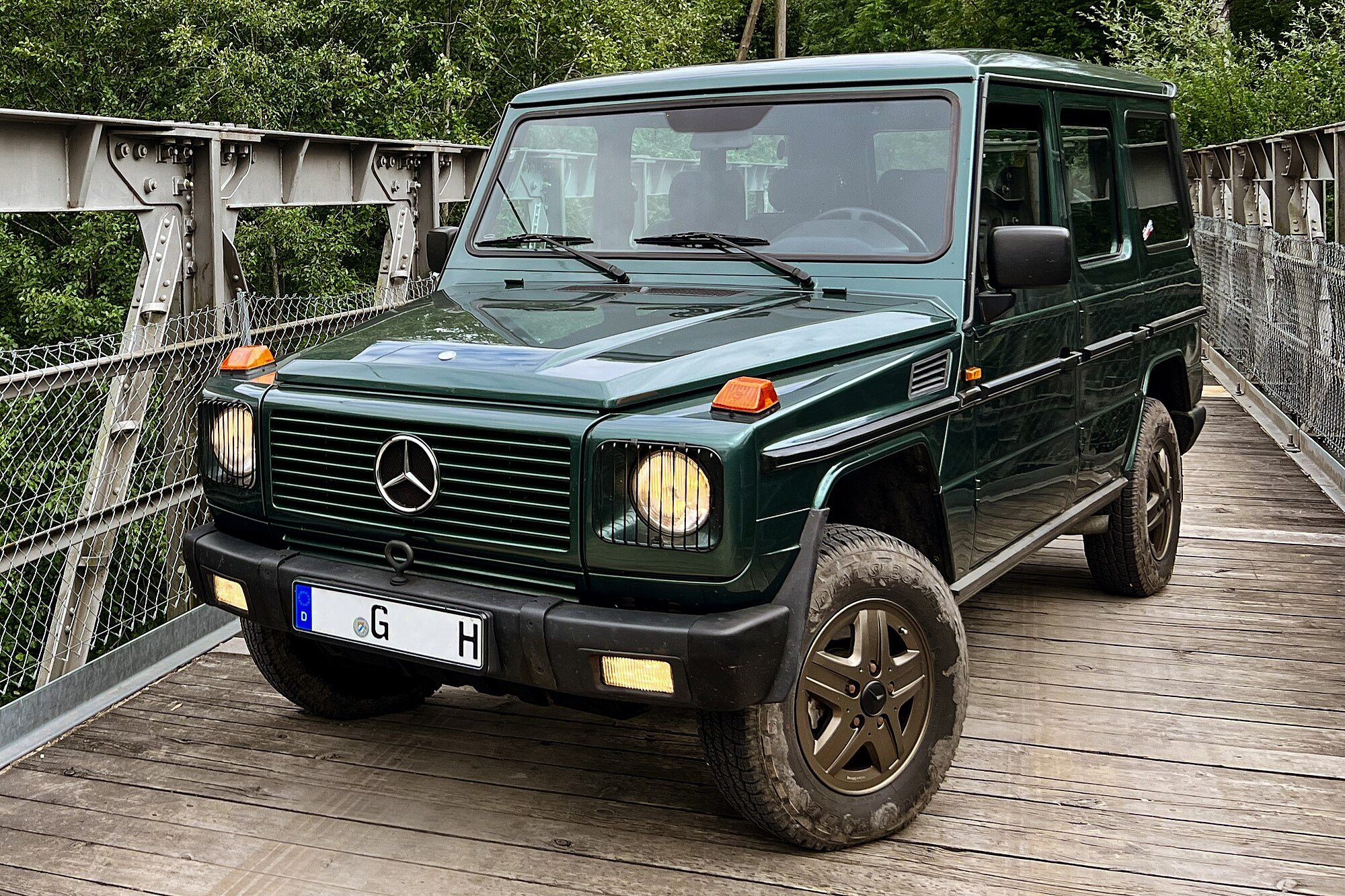 A dark green early-1990s W463 300 GD long-wheelbase on the timber deck of a steel footbridge among trees