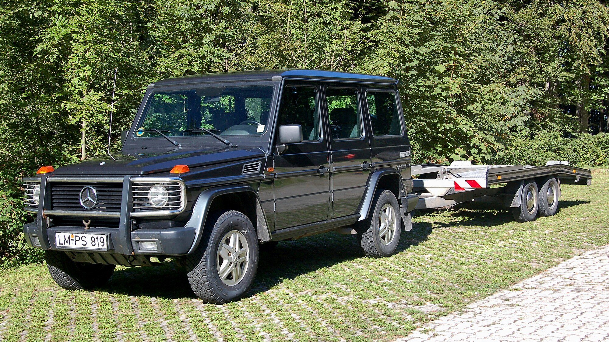 A dark blue W463 long-wheelbase with bull bar parked on grass-block paving with a car trailer, woods behind