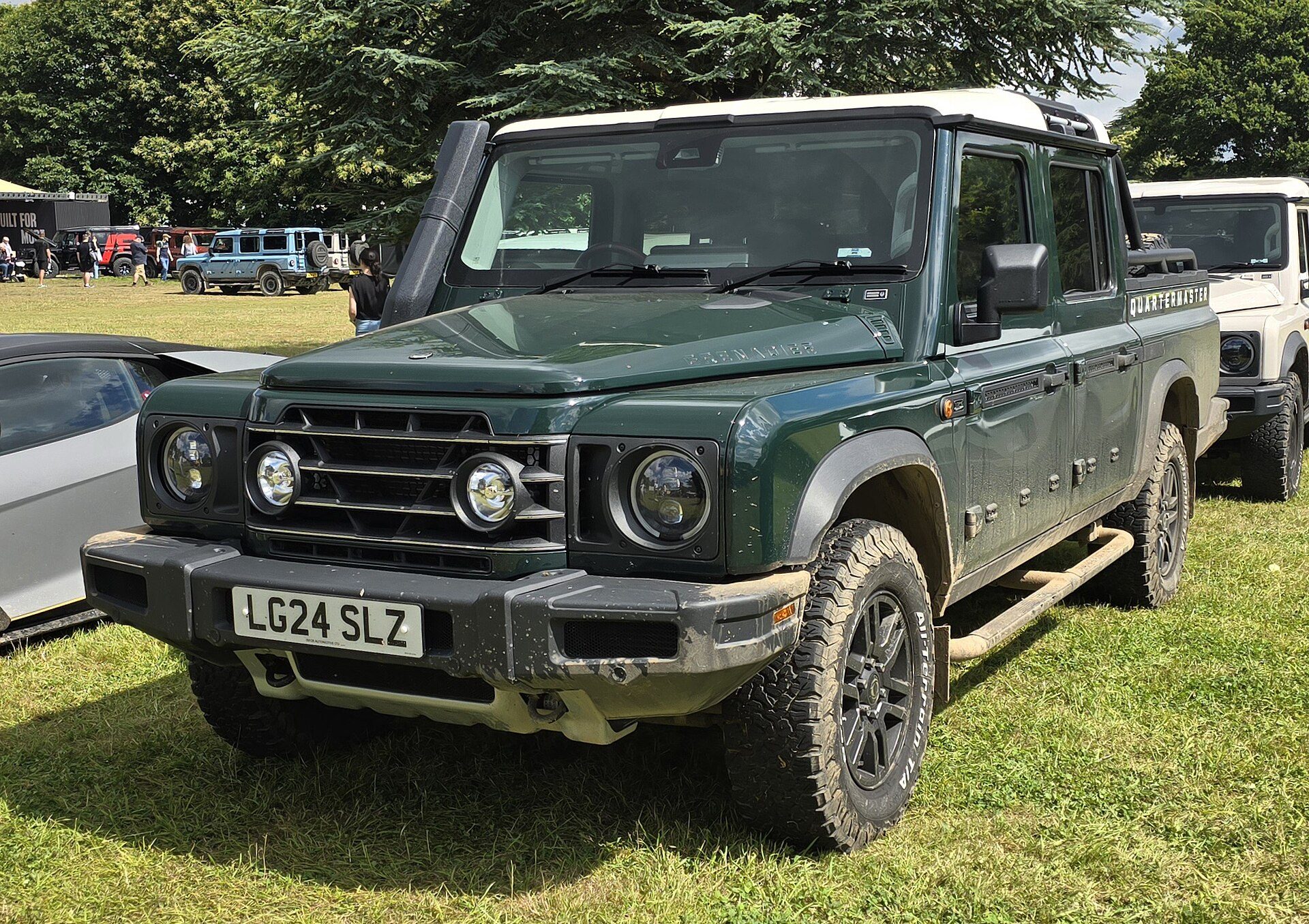 A dark green Ineos Grenadier Quartermaster Trailmaster Edition with snorkel and muddy bumper, front three-quarter view on grass