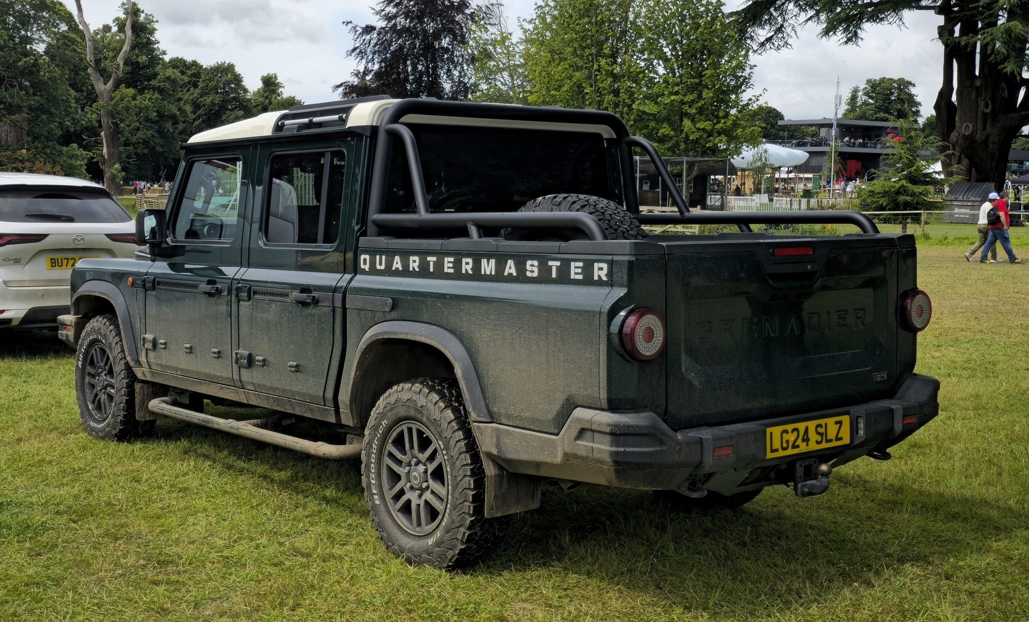 A dark green Ineos Grenadier Quartermaster double-cab pickup with sports bar and spare wheel in the bed, rear three-quarter view on grass