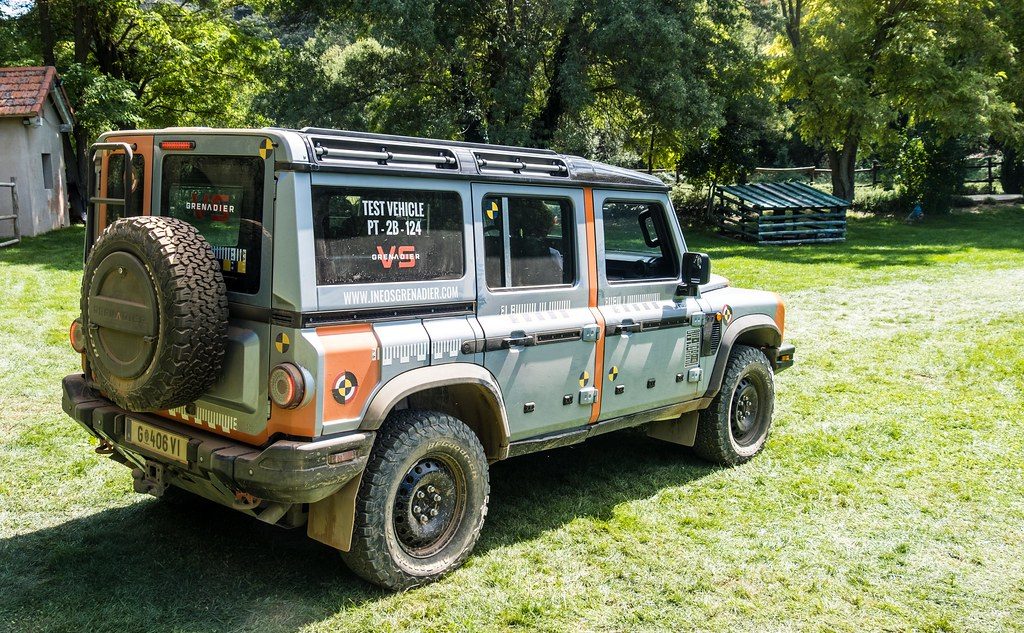 A muddy, grey-and-orange Ineos Grenadier Station Wagon test vehicle with a rear ladder and roof rack, parked in a rural meadow beside farm buildings