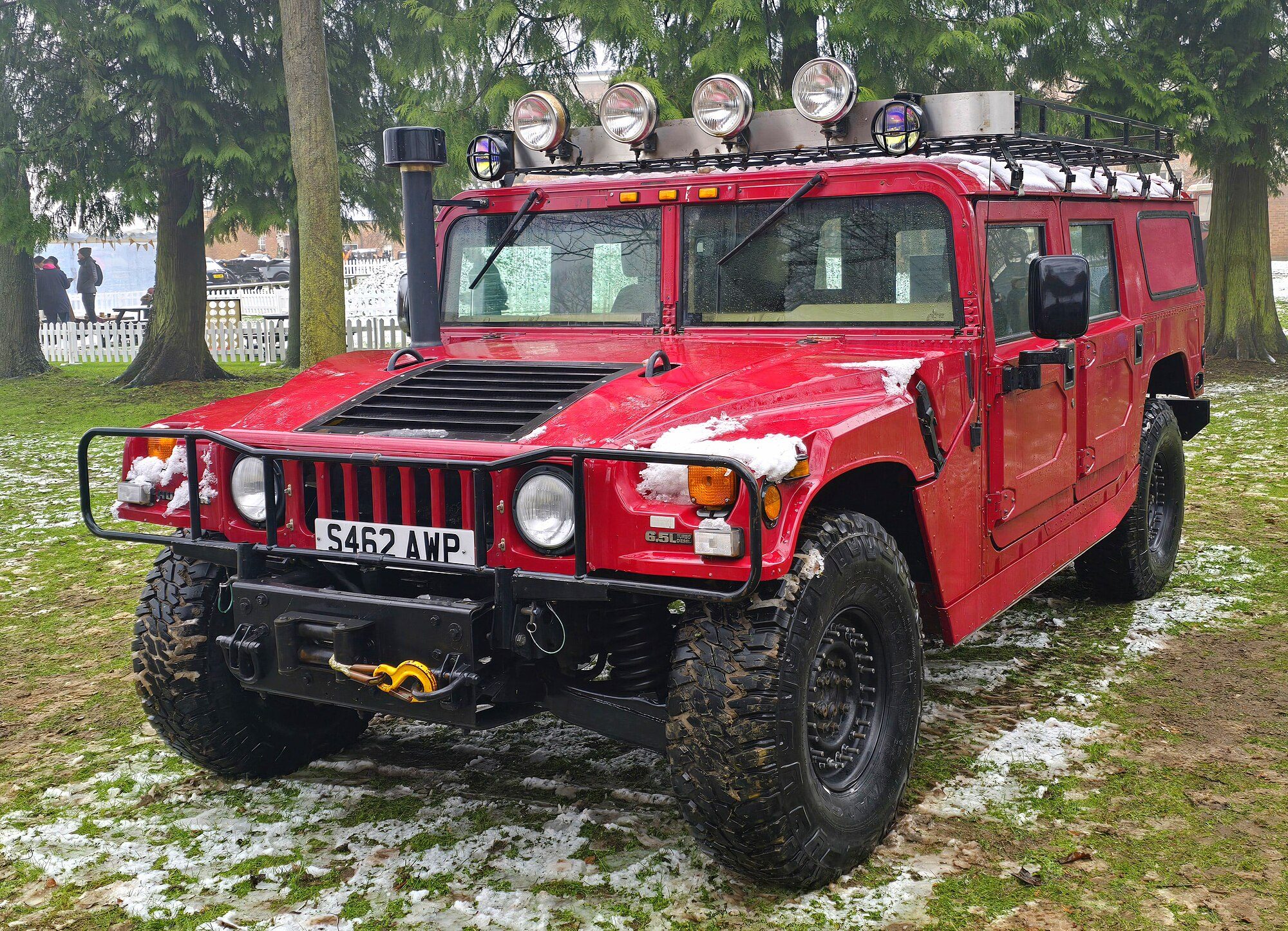 A red Hummer H1 wagon with roof lights, snorkel and winch parked on snow-dusted grass among fir trees
