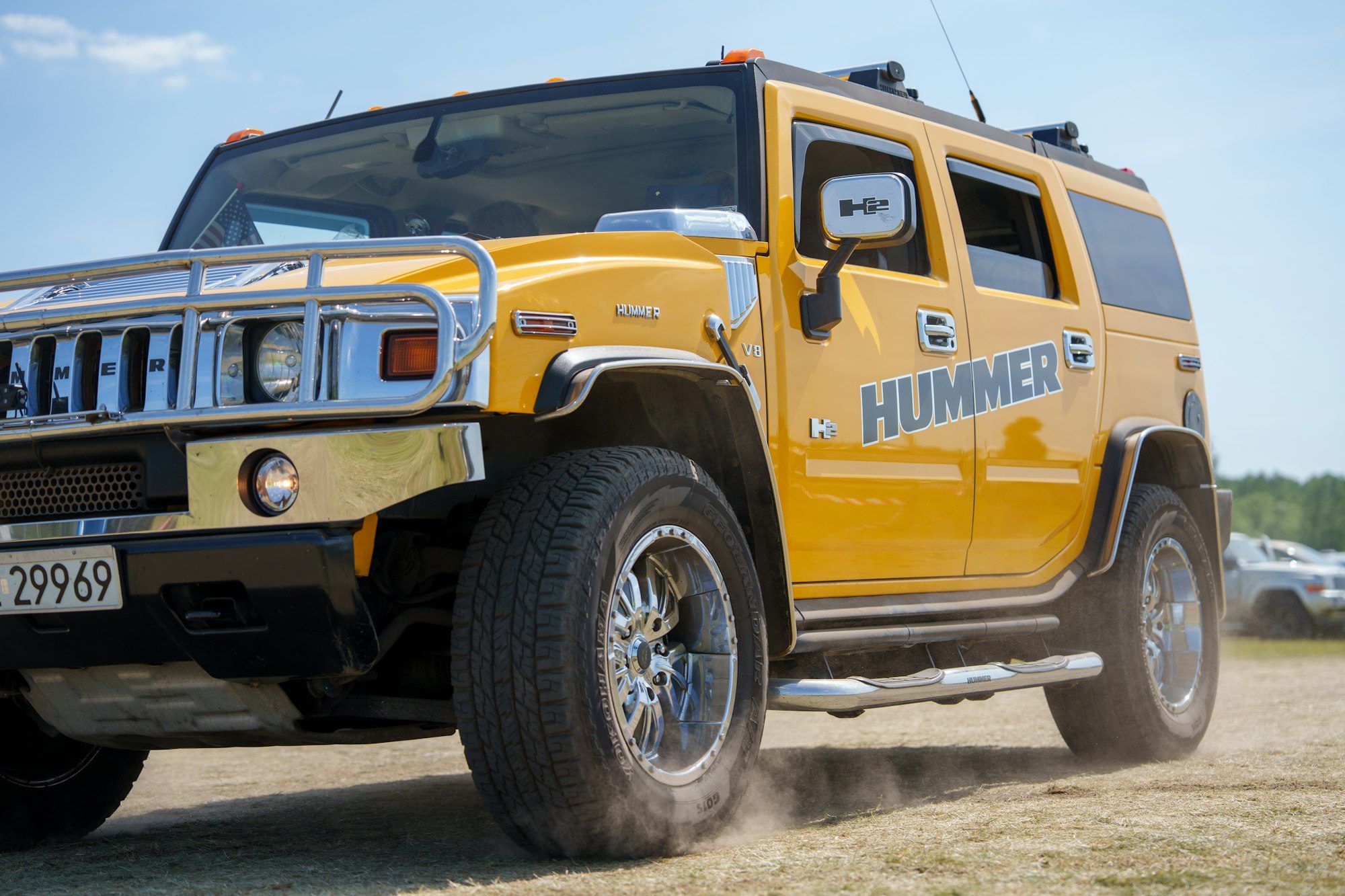 A yellow Hummer H2 with a chrome brush guard and HUMMER door graphics kicking up dust on a dry dirt field