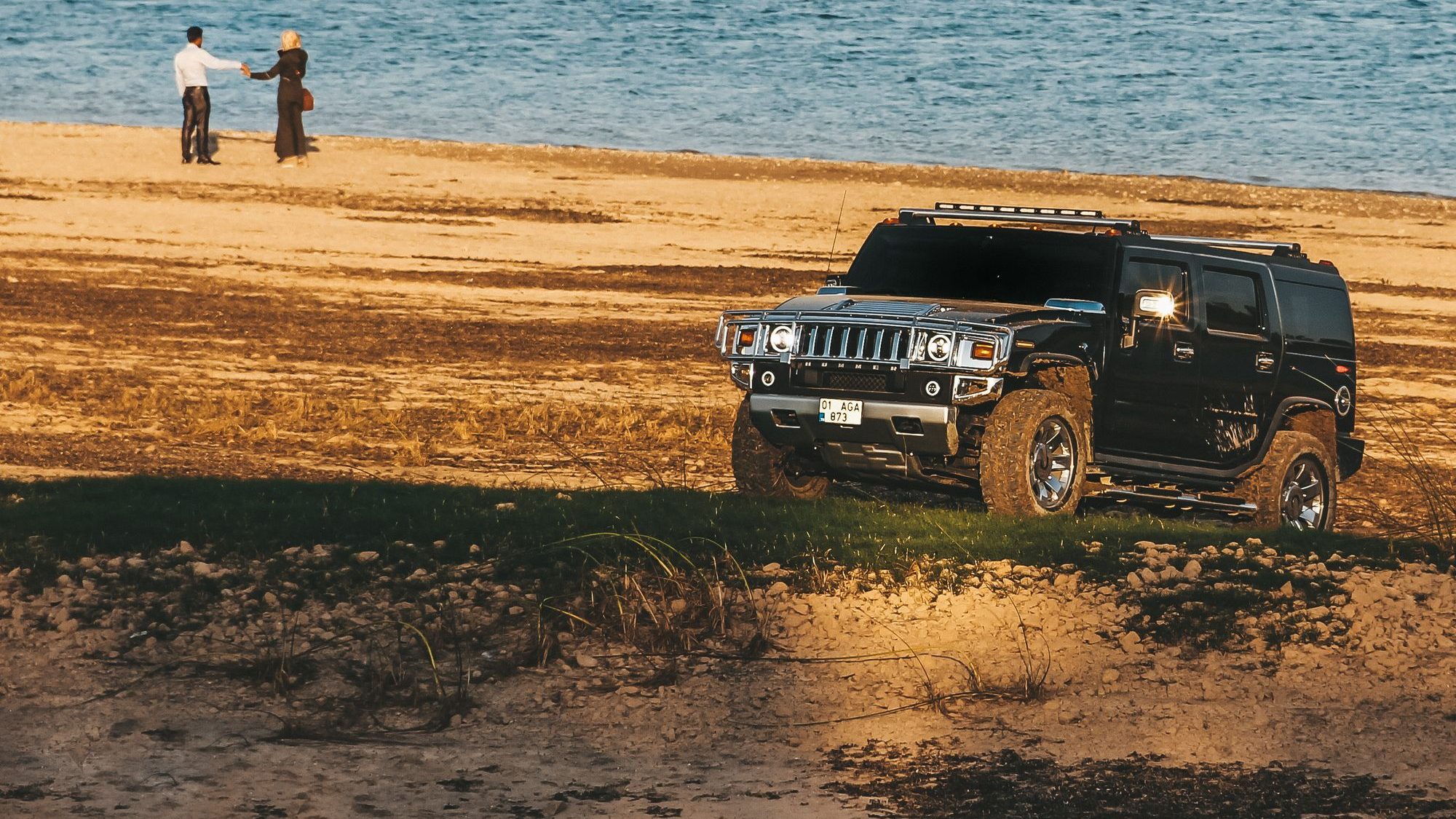 A black Hummer H2 with a roof rack parked on a grassy rise above a sandy lakeshore, two people walking by the water behind it