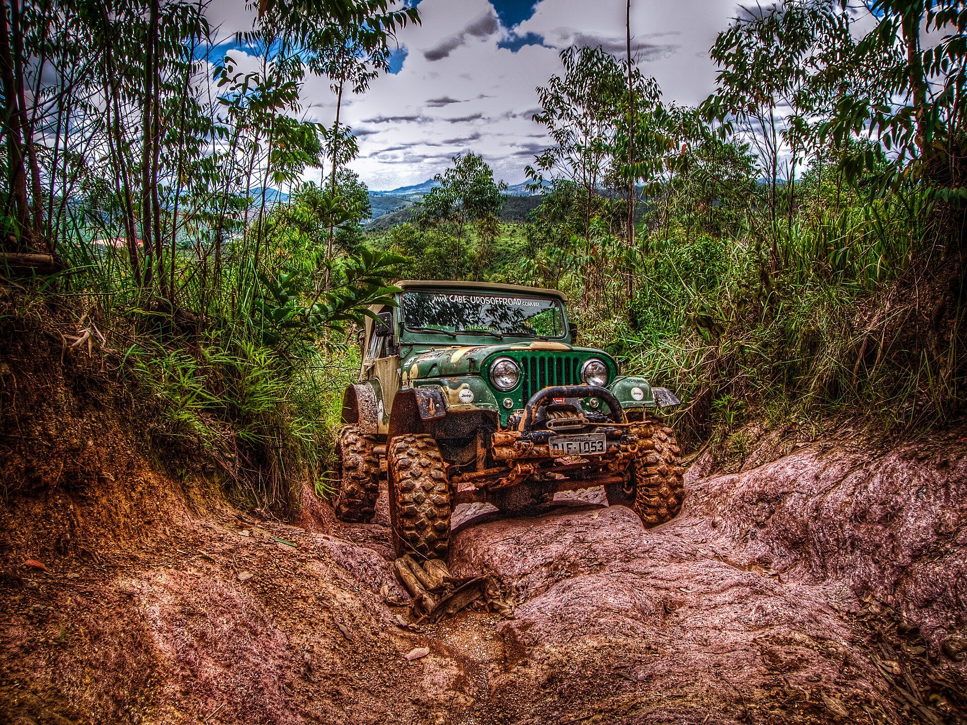 The same lifted green CJ-5-type Jeep climbing out of a deep red-clay gully between banks of grass and trees