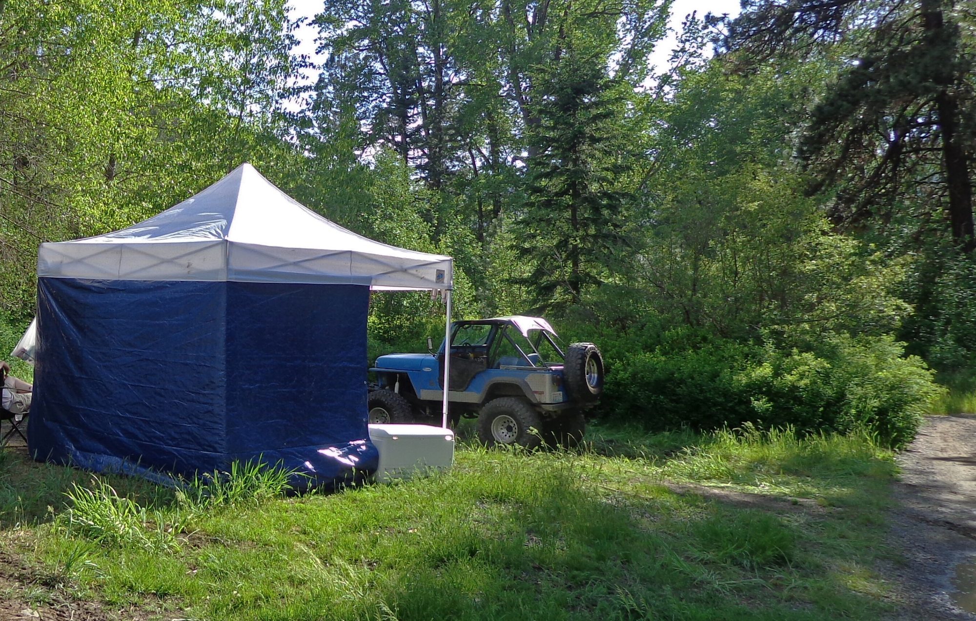 A lifted blue Jeep CJ-5 with a bikini top, roll bar and big mud tires parked beside a canopy at a forest campsite in the Oregon Cascades