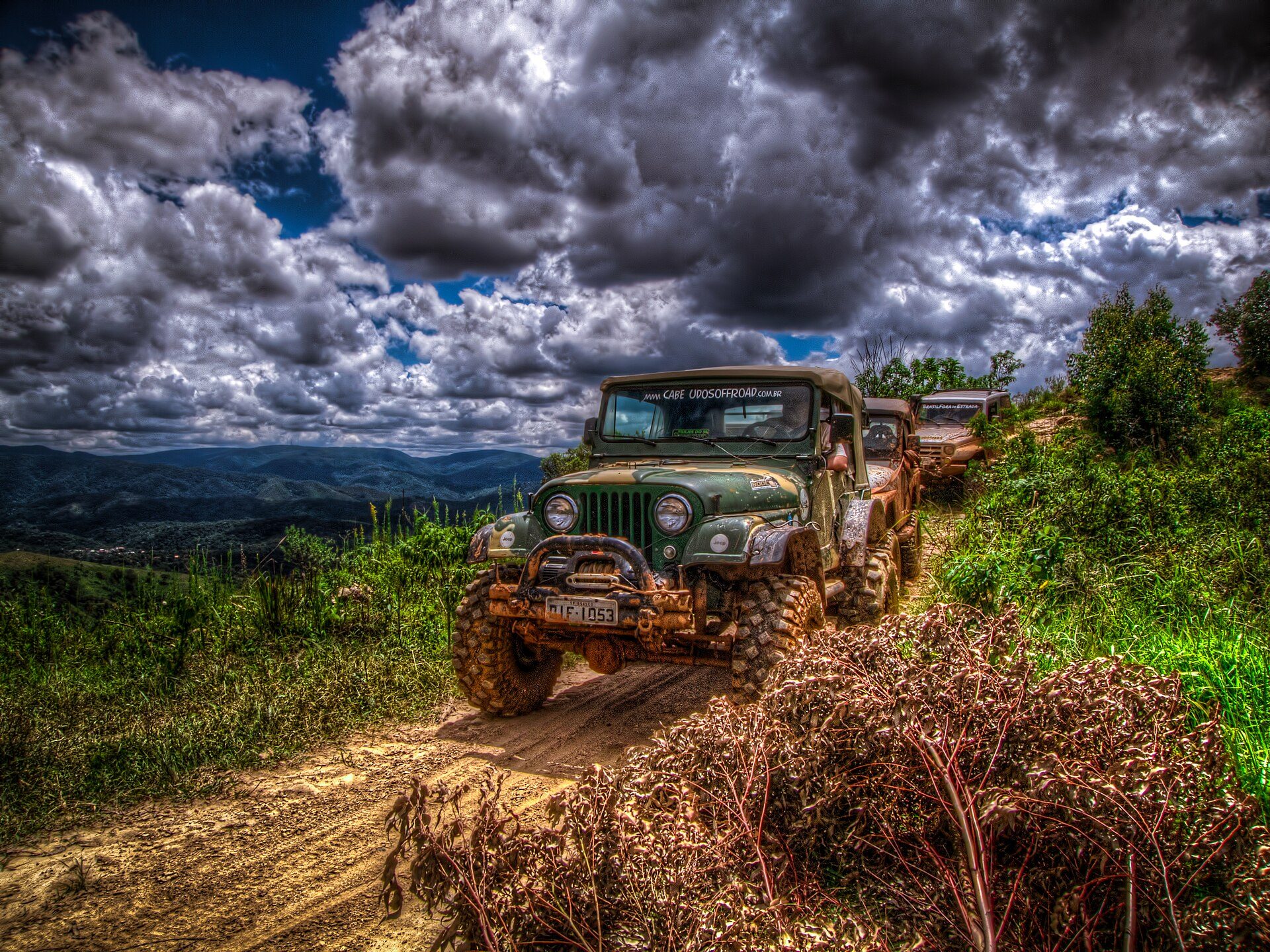 A green, Brazilian-registered CJ-5-type Jeep on oversized mud tires leading a line of Jeeps along a dirt trail above a valley in Brazil