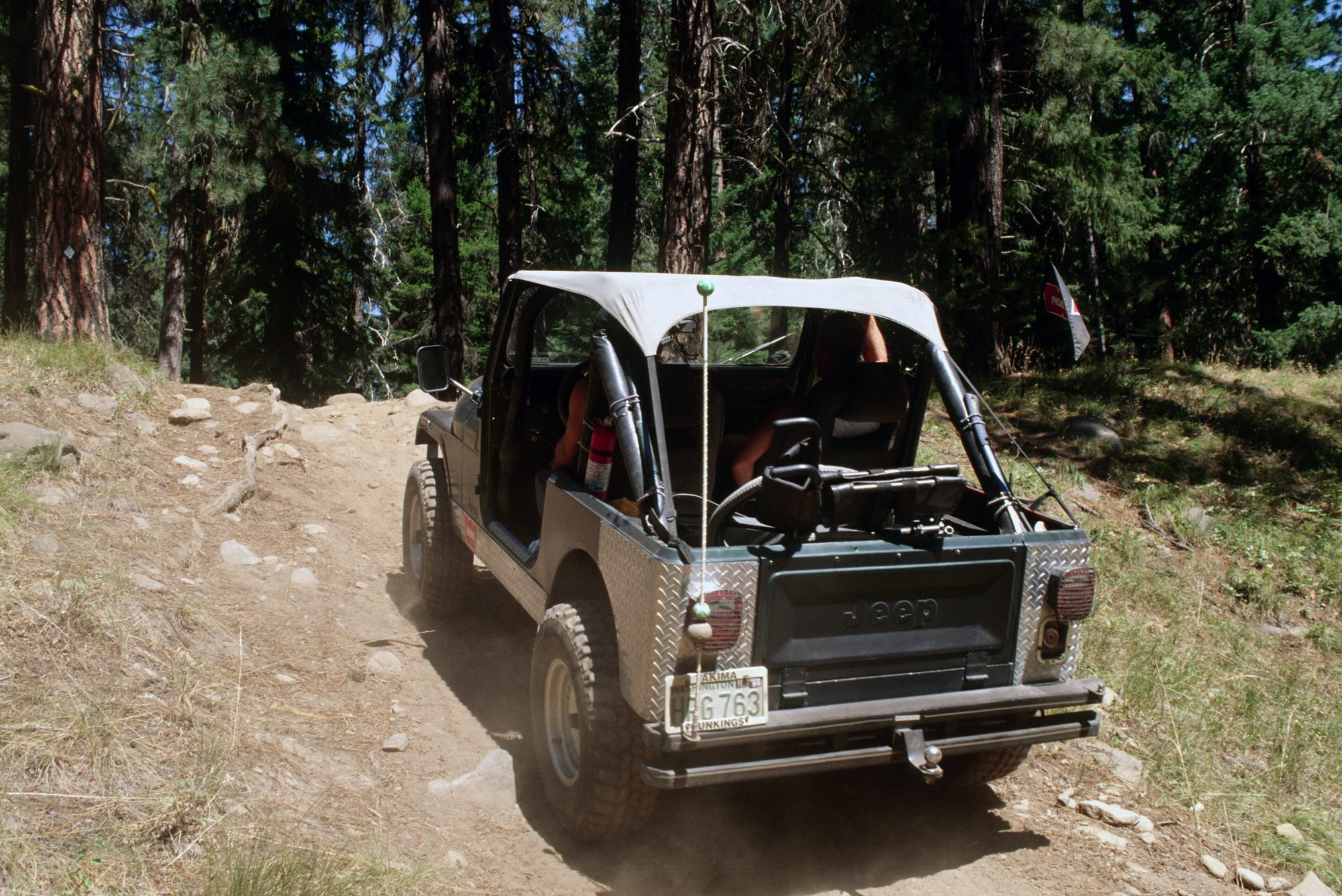 A CJ-7 with a bikini top and diamond-plate corners climbing a dusty forest trail