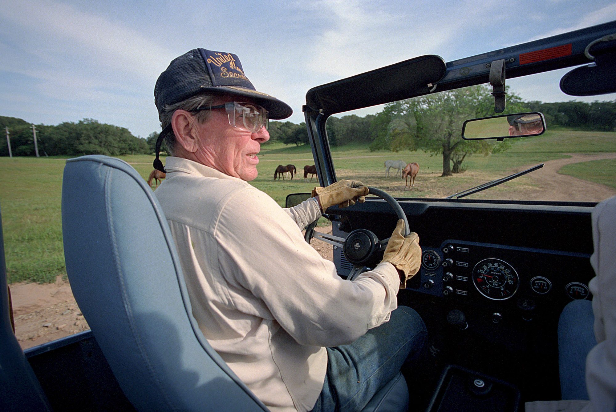 Ronald Reagan in work gloves at the wheel of his open-topped blue Scrambler, with horses grazing in the ranch pasture behind