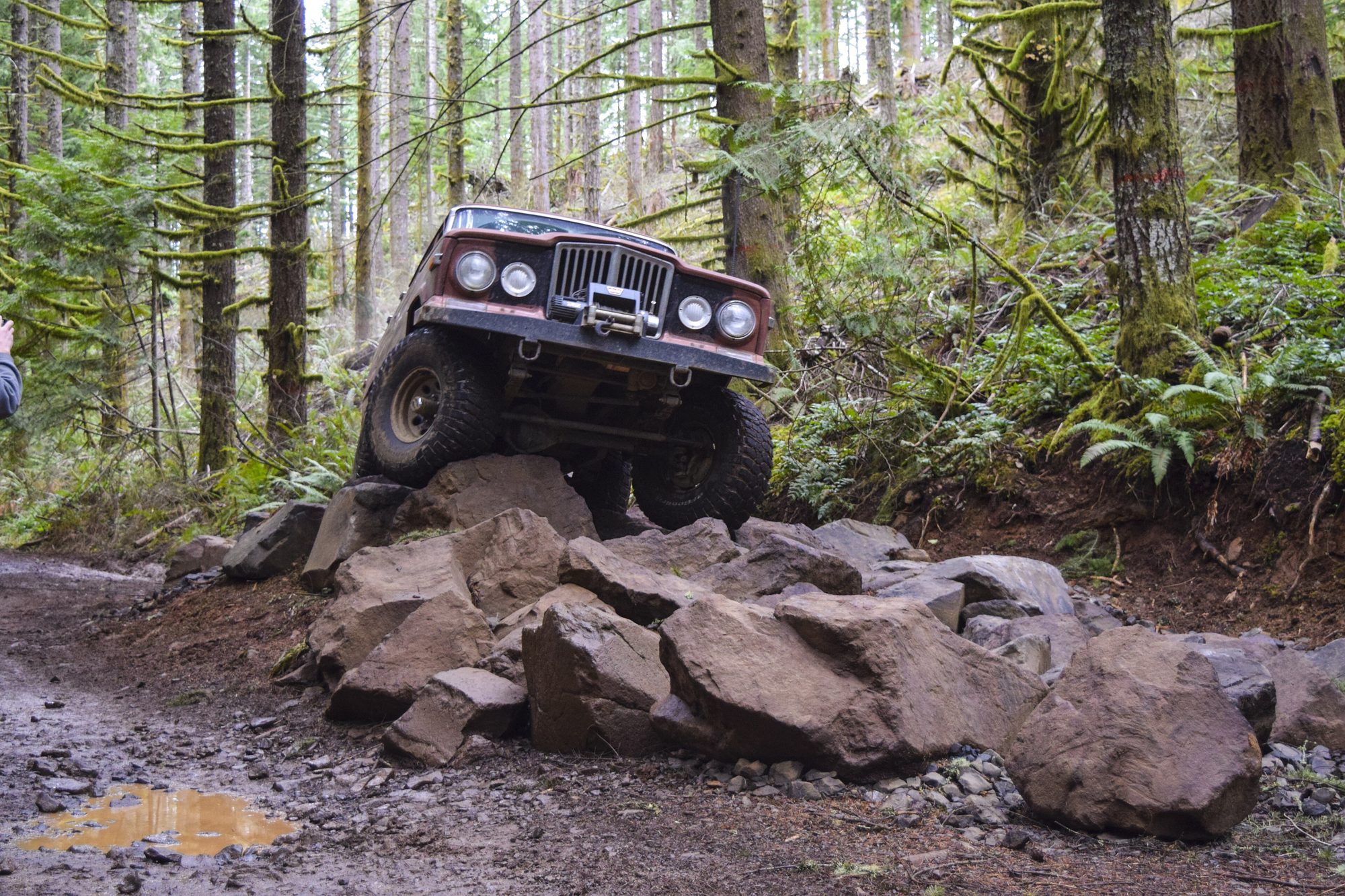 A heavily modified full-size Jeep SJ crawling over a pile of basalt boulders on a muddy forest trail