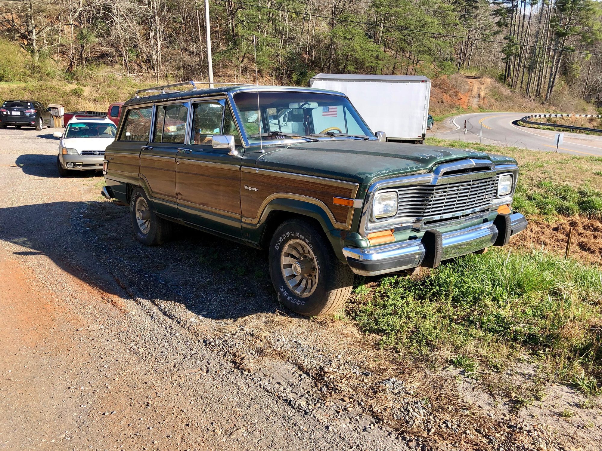A dark green 1986–91 Jeep Grand Wagoneer with woodgrain sides parked on a red-dirt gravel pull-off beside a mountain road in western North Carolina