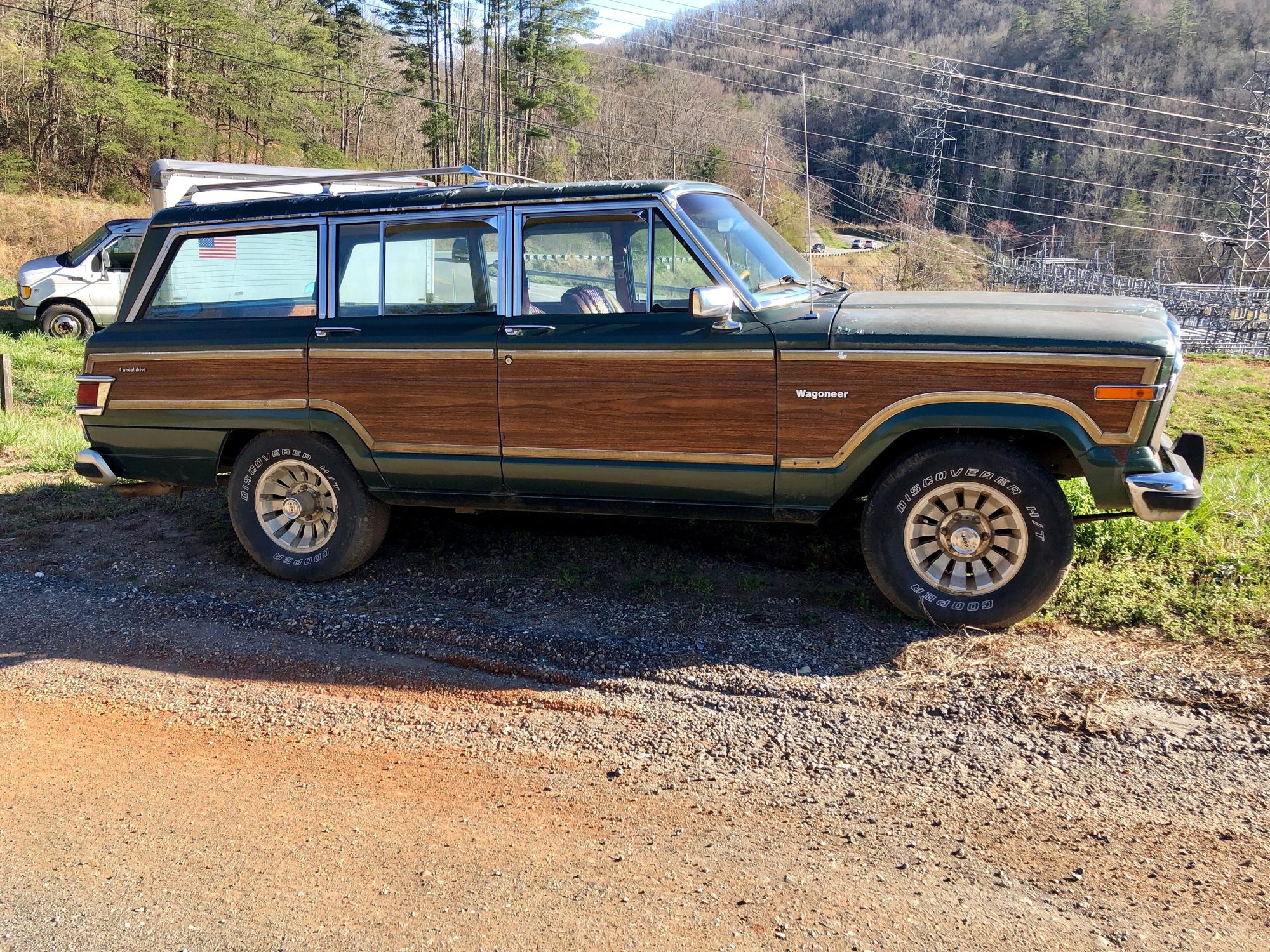 Side view of a green Grand Wagoneer with full-length woodgrain trim on a gravel pull-off beside a wooded hillside