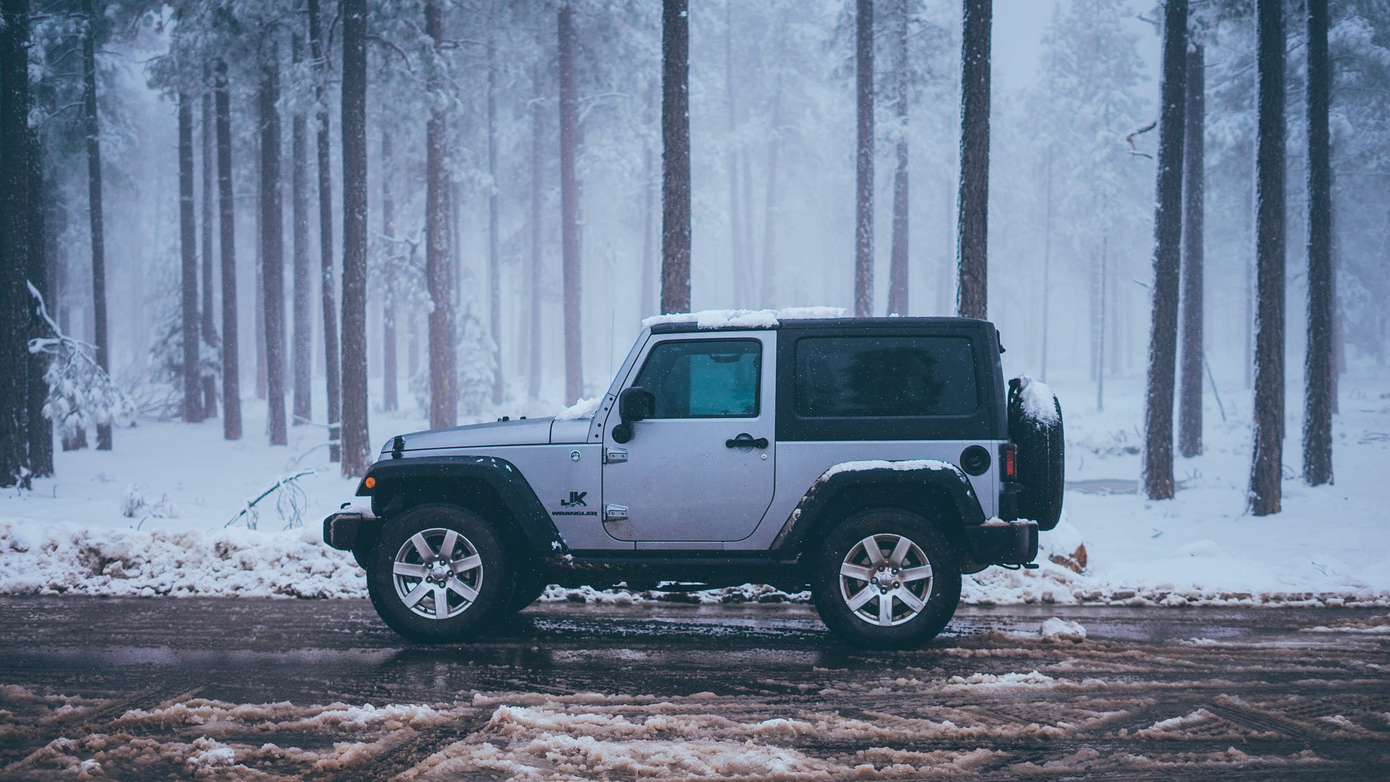 A silver two-door Jeep Wrangler JK with a 'JK' fender decal on a wet, slushy road through a misty, snowy pine forest