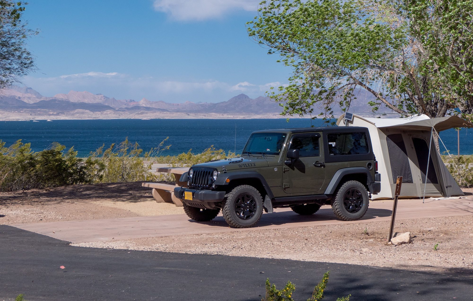 A dark green two-door Wrangler JK Willys Wheeler parked at a lakeside campsite beside a tent, with Lake Mead and desert mountains behind