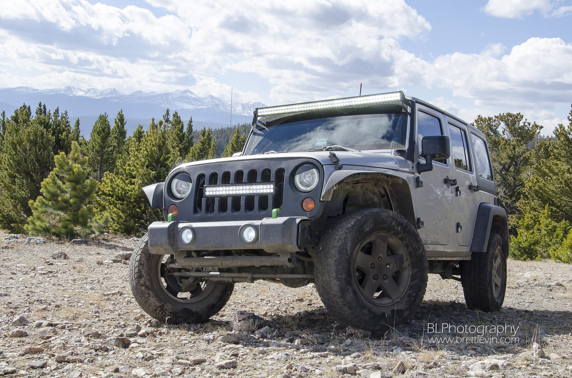 A grey lifted Jeep Wrangler JK Unlimited on a rocky gravel summit with pine forest and snow-capped mountains behind