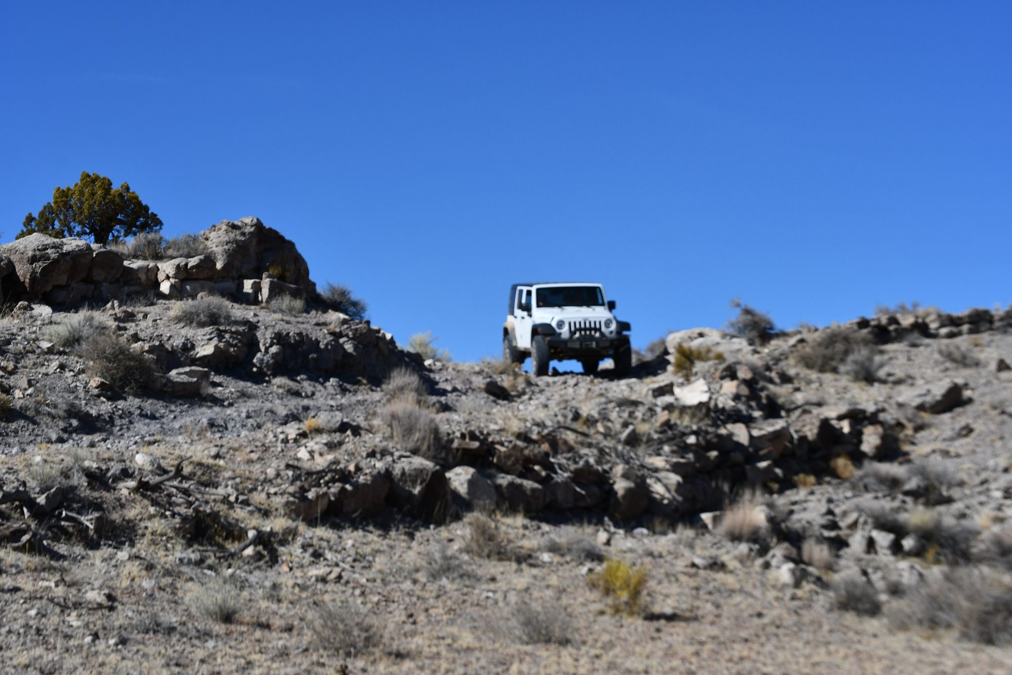 A white Jeep Wrangler JK, seen head-on, cresting a rocky dugway under a deep blue sky in Emery County, Utah